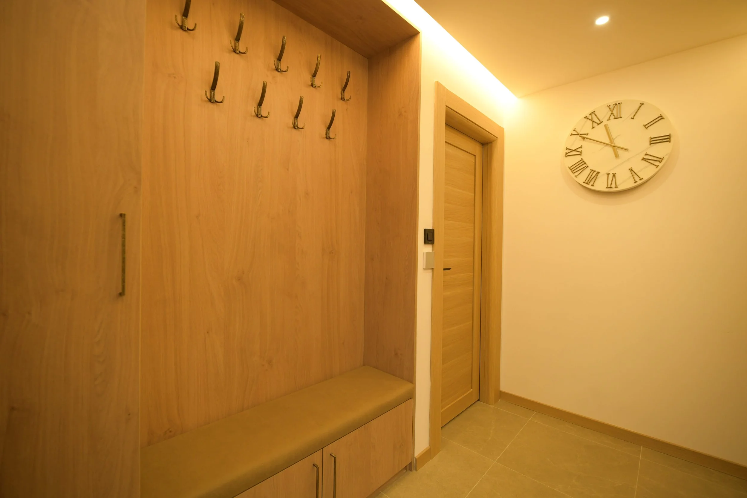 An entryway with a wooden wall featuring hooks for hanging items, a bench below the hooks, a closed wooden door, and a large wall clock showing the time as 10:11.