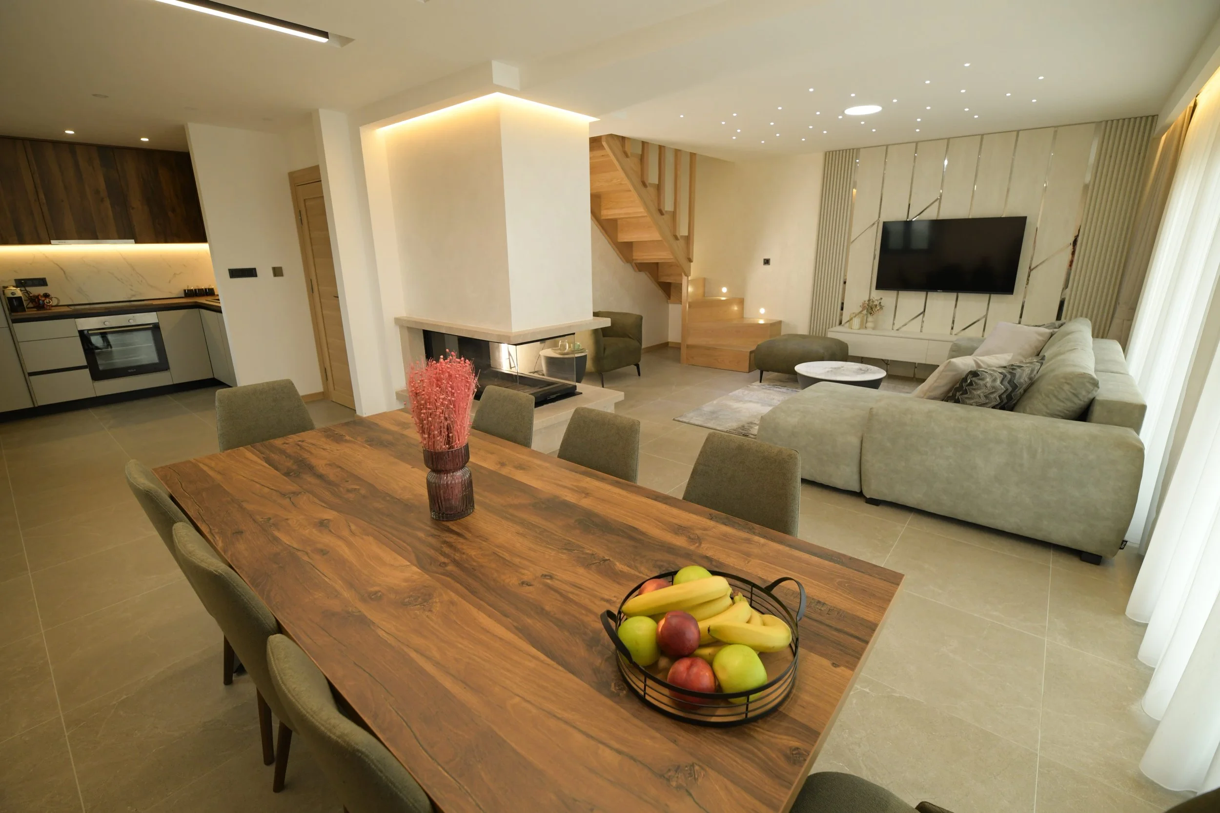 Open-concept living and dining area with wooden dining table, chairs, a modern kitchen, a sectional sofa, a wall-mounted TV, a fireplace, and a staircase with wooden steps.