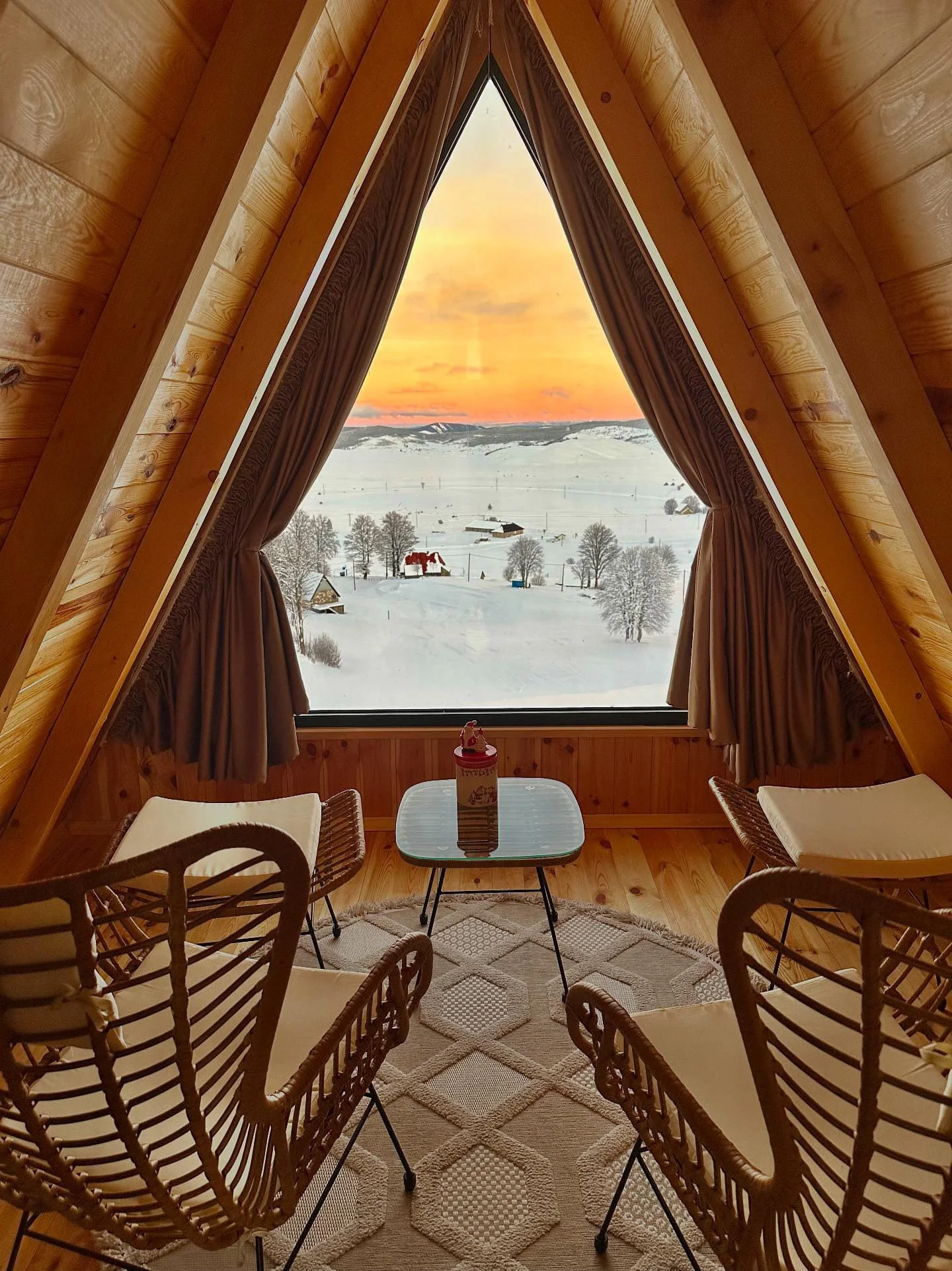 A cozy attic room with wooden walls and ceiling, featuring a large triangular window with brown curtains, overlooking a snowy landscape with trees and distant mountains at sunset. There are four wicker chairs with cushions arranged around a small whi