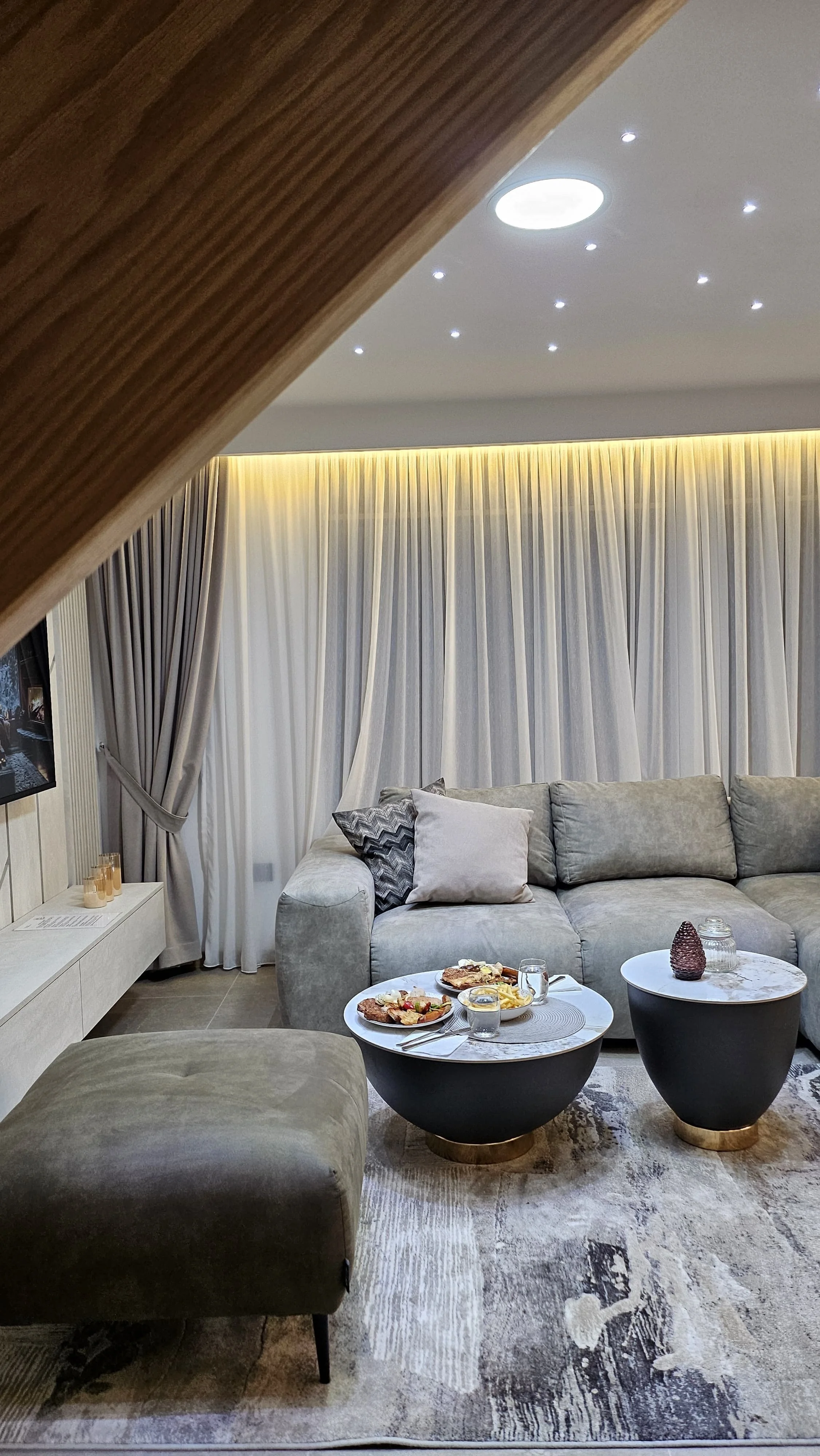 Living room with a grey sectional sofa, round coffee tables with food, and beige curtains.