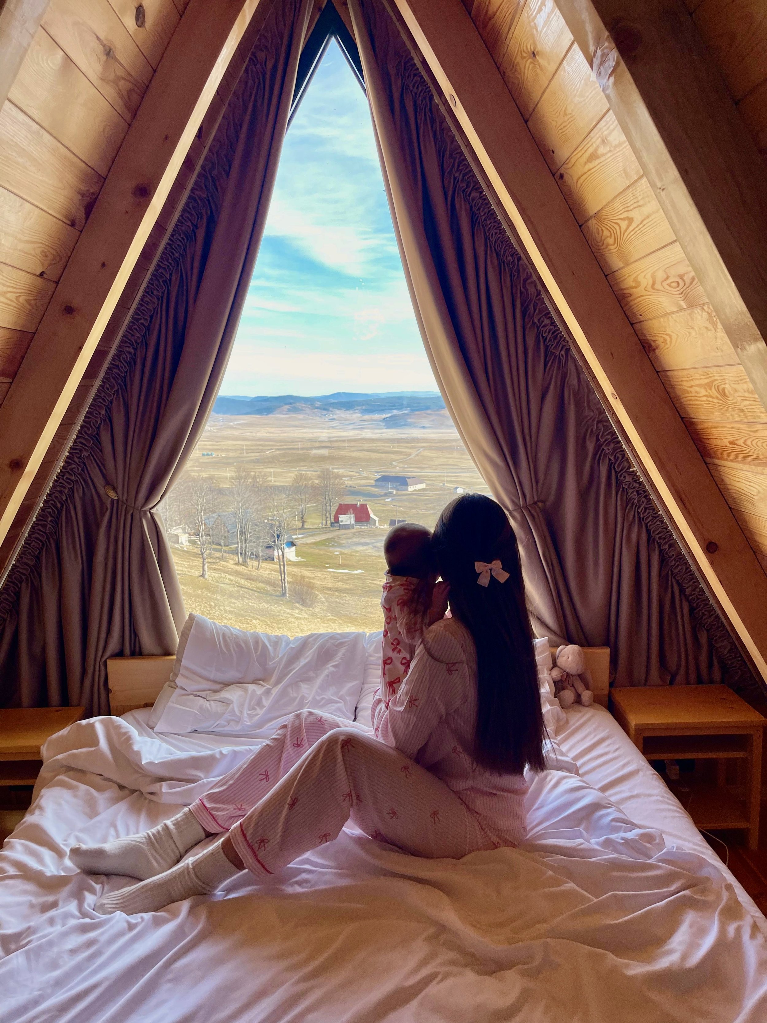 A girl in pink pajamas with a bow in her hair, sitting on a bed with white sheets, holding a small stuffed animal, in a cozy wooden attic room with large triangular window overlooking a rural landscape.