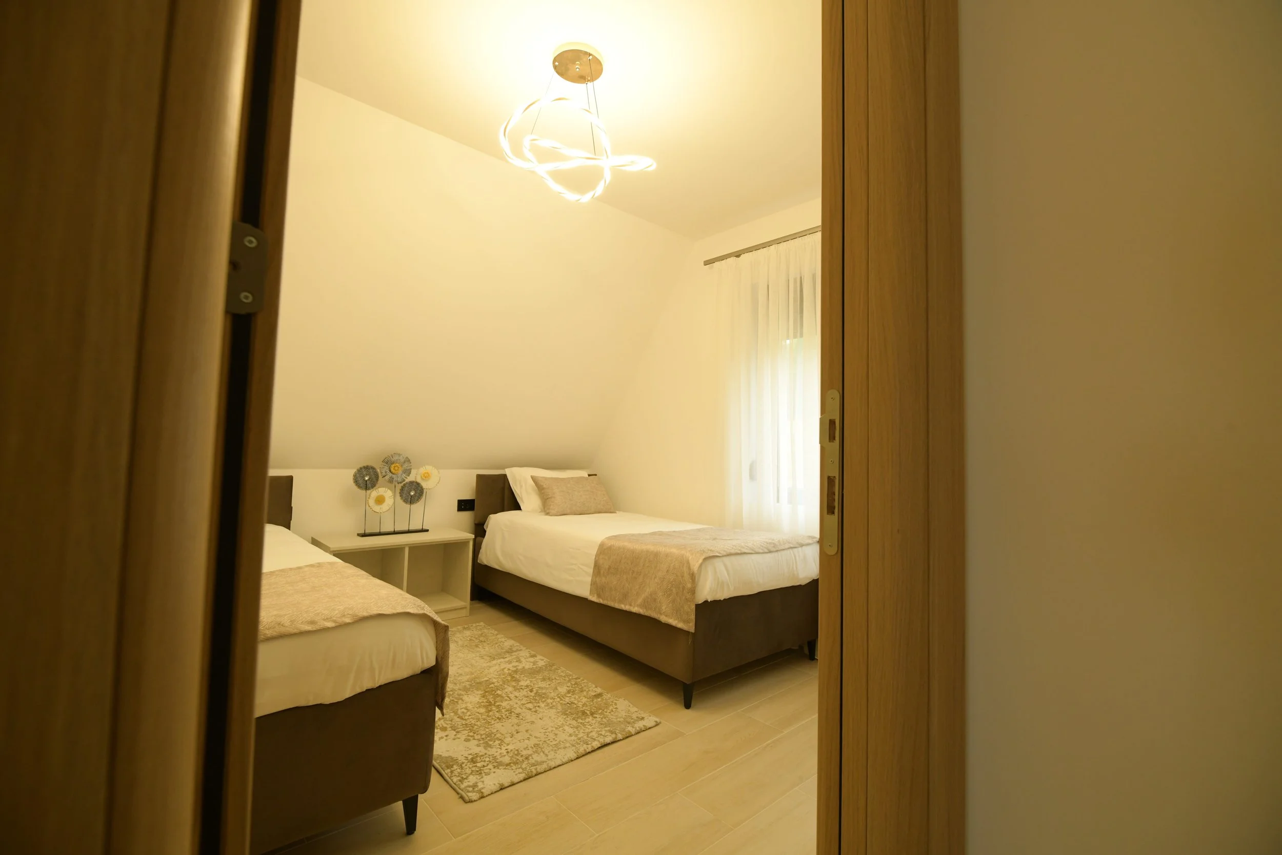 View of a modern bedroom with two beds, decorative flowers on a white nightstand, beige curtains, a rug, and a ceiling light fixture, seen through a partially opened wooden door.