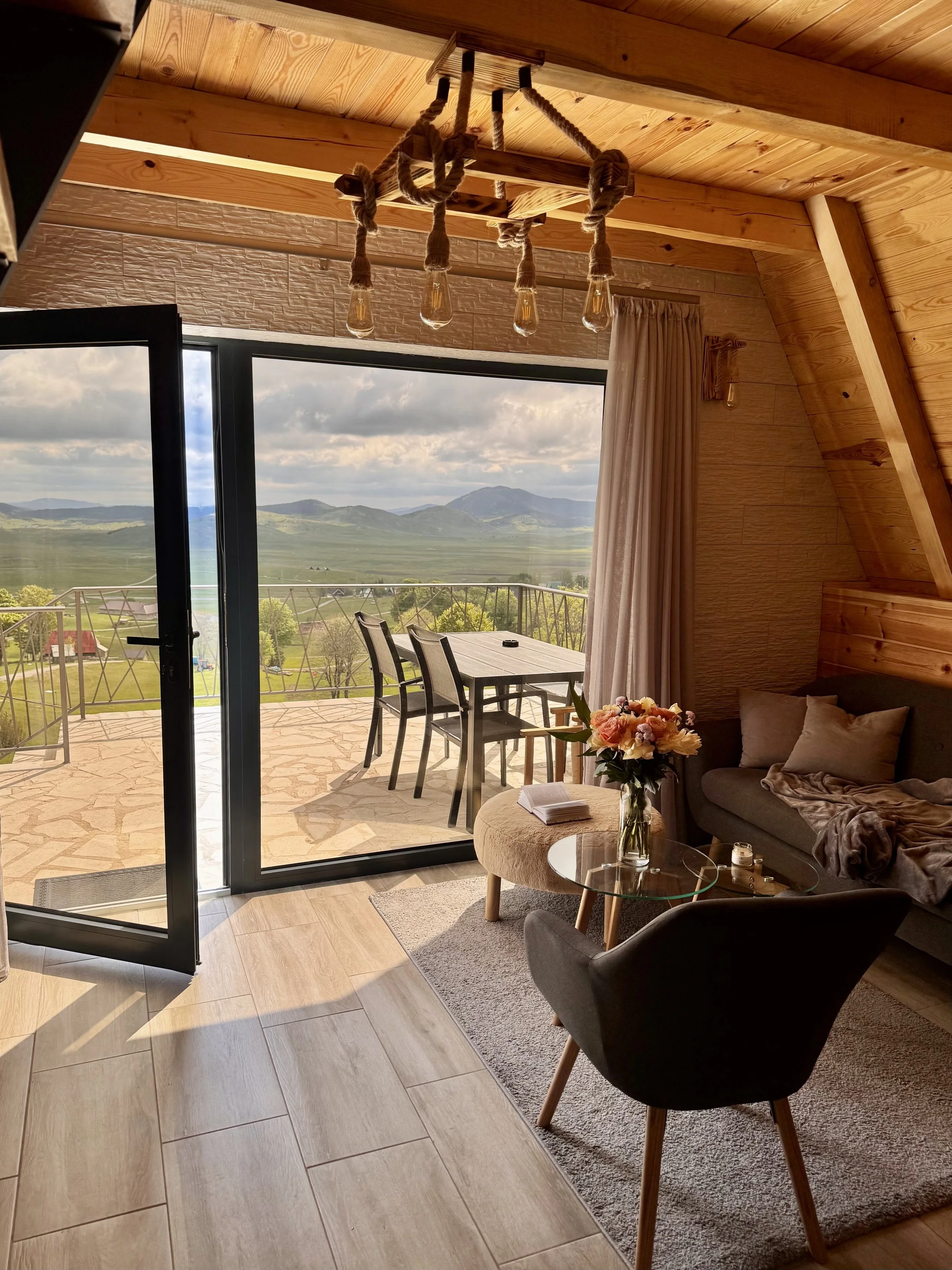 A cozy living room with wooden walls and ceiling, featuring a sliding glass door open to a balcony. The balcony has outdoor furniture and offers a scenic view of rolling green hills and mountains in the distance. Inside, a sofa with cushions, a small