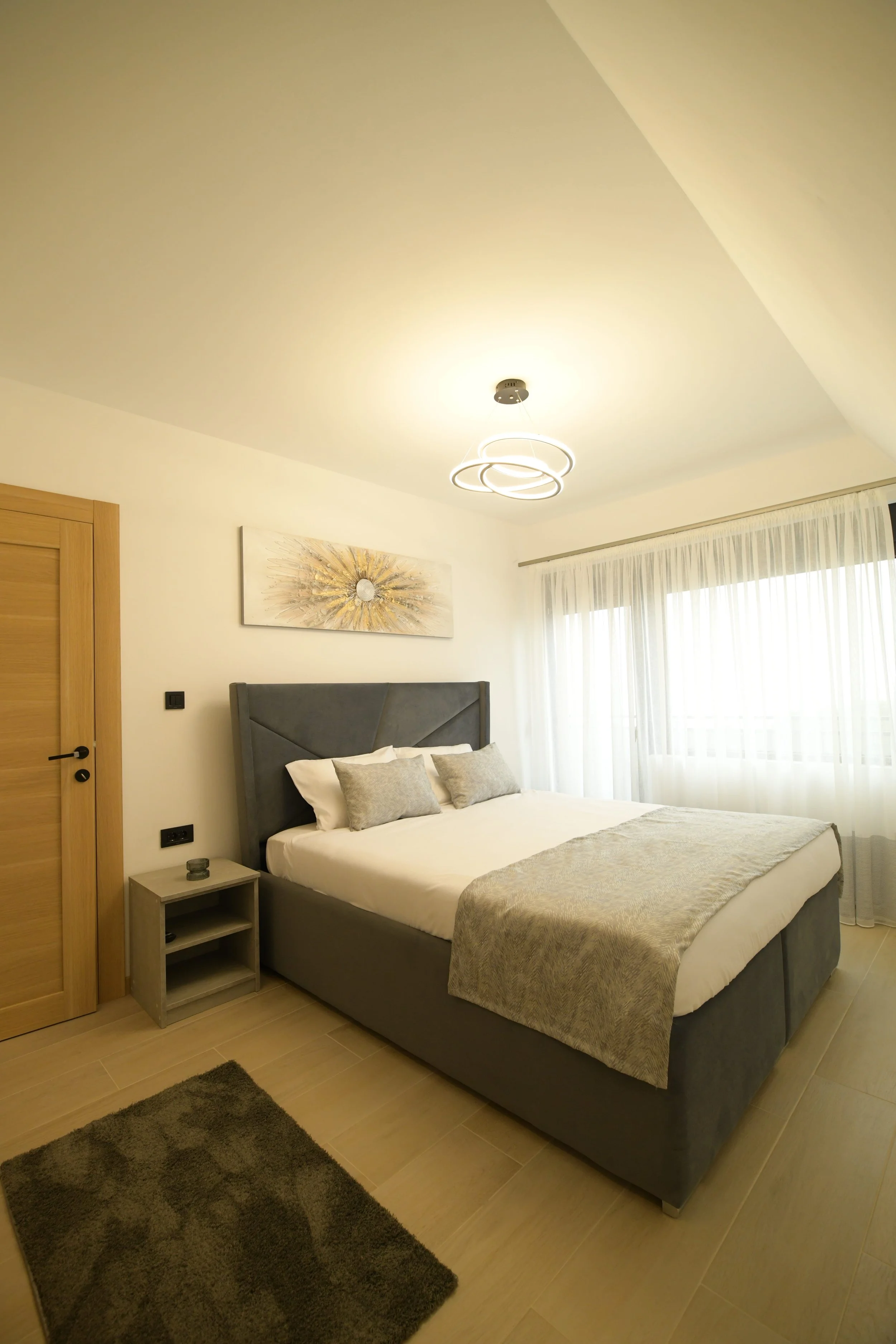 A modern, well-lit bedroom with a large bed, grey upholstered headboard, decorative pillows, cream-colored bedspread, wooden bedside table, abstract wall art, window with sheer curtains, and a ceiling light fixture.