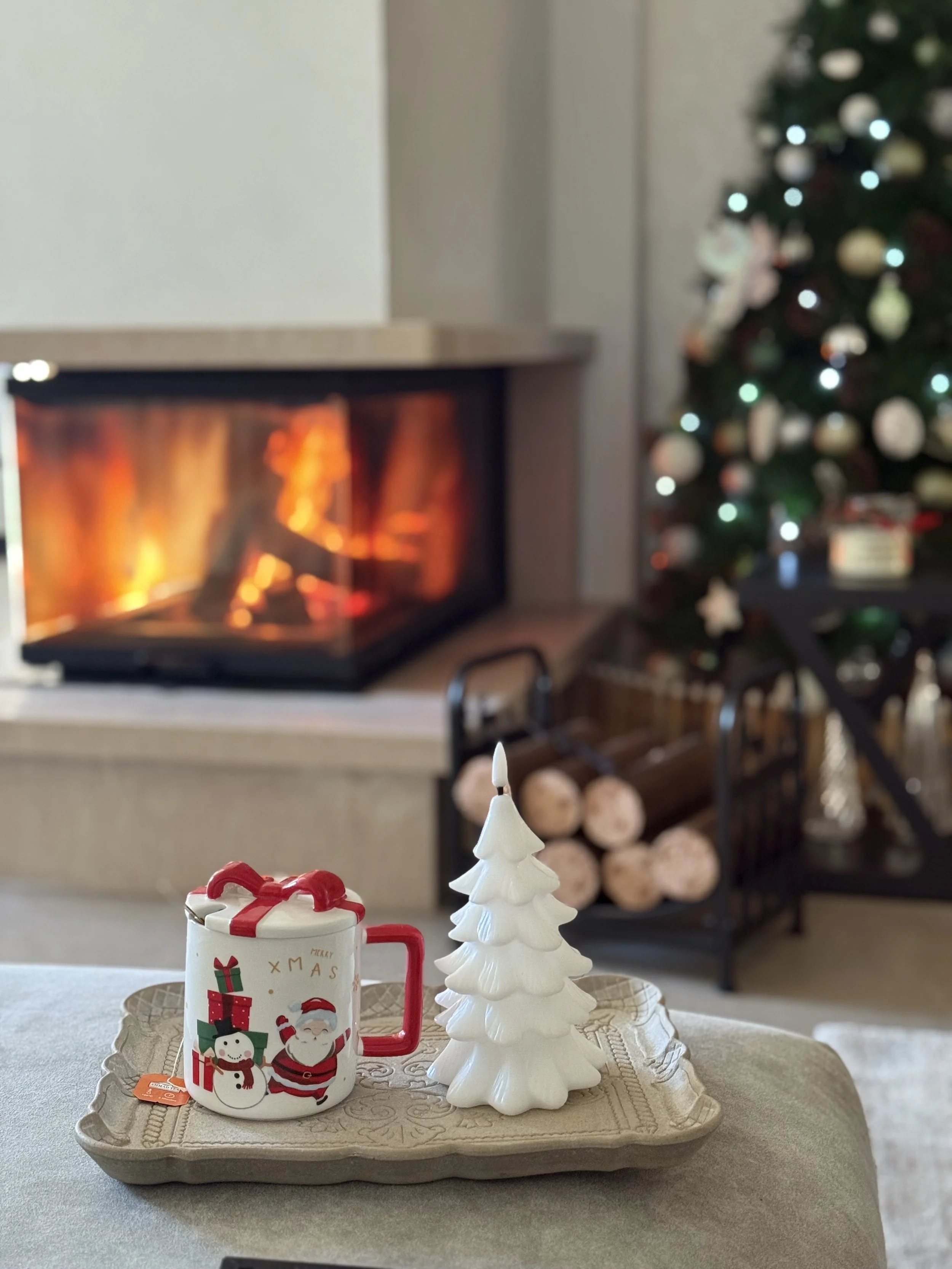 Christmas themed tray with a mug decorated with a snowman and Santa, and a white ceramic Christmas tree, placed in front of a lit fireplace and a decorated Christmas tree in the background.