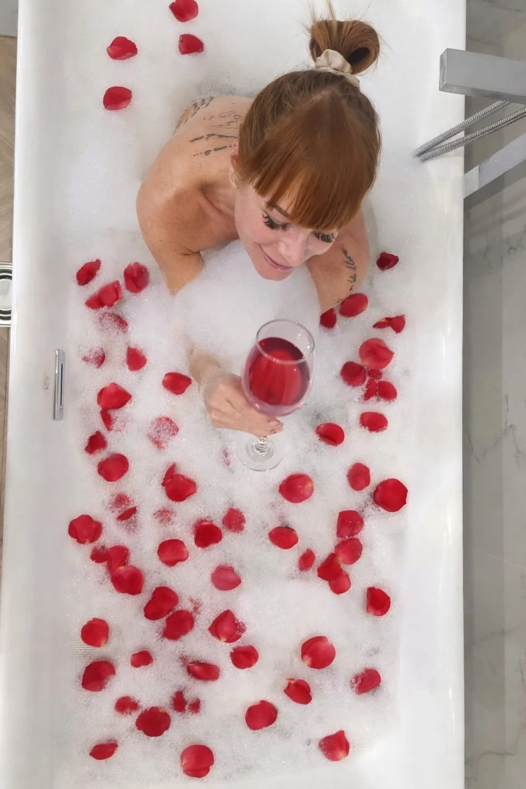 A woman with red hair and tattoos relaxing in a bubble bath filled with red rose petals, holding a glass of red wine.