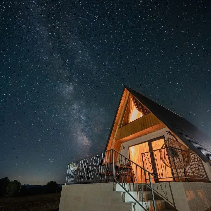 Aerial view of a modern A-frame house with an illuminated interior, balcony, and stairs, set against a starry night sky with visible Milky Way galaxy.