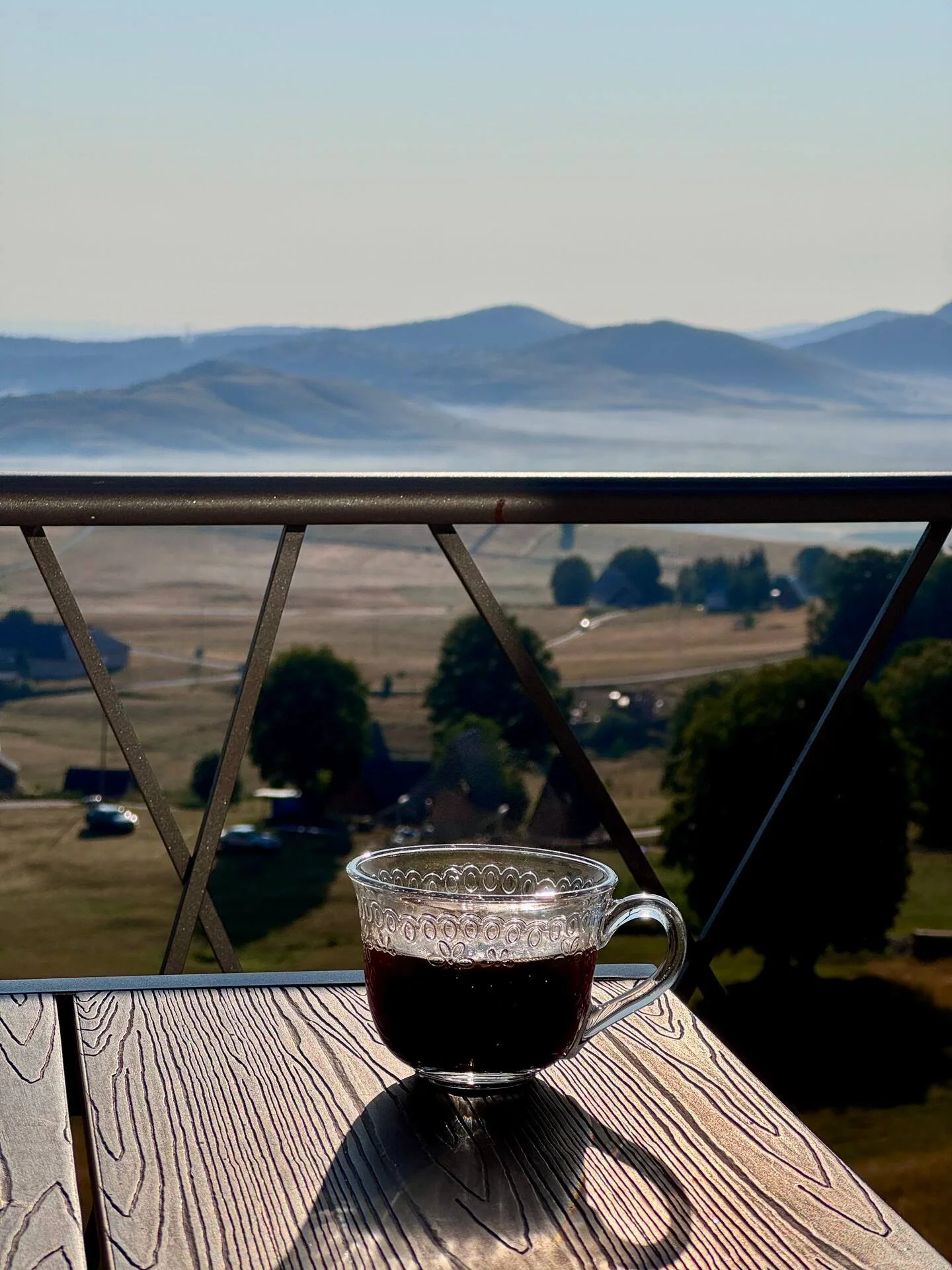 A glass cup filled with dark coffee on a wooden table, with a scenic background of mountains, trees, and a rural landscape.
