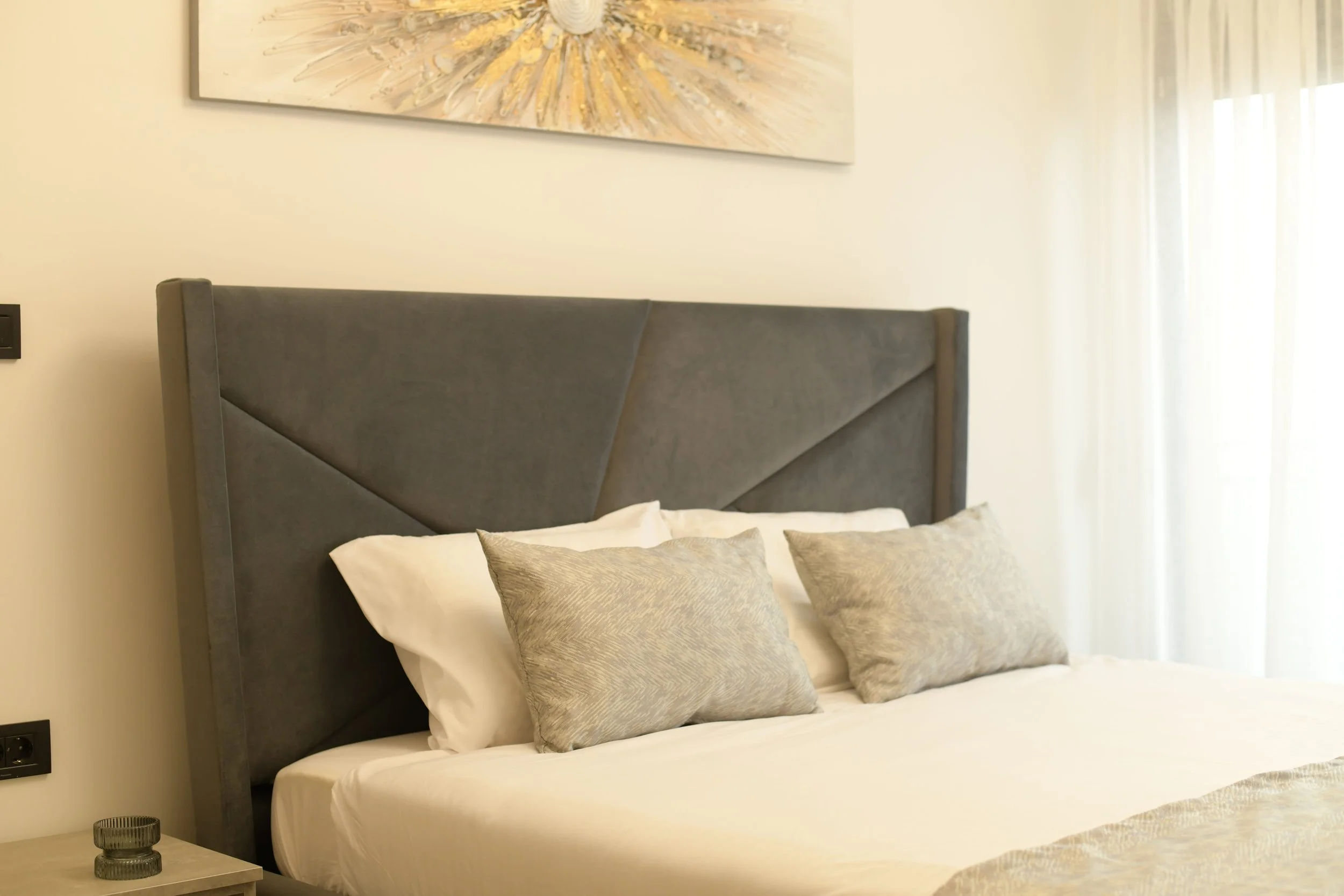 A neatly made bed with a gray upholstered headboard and three textured gray pillows, situated in a modern, well-lit bedroom with a cream-colored wall and a large abstract artwork hanging above.