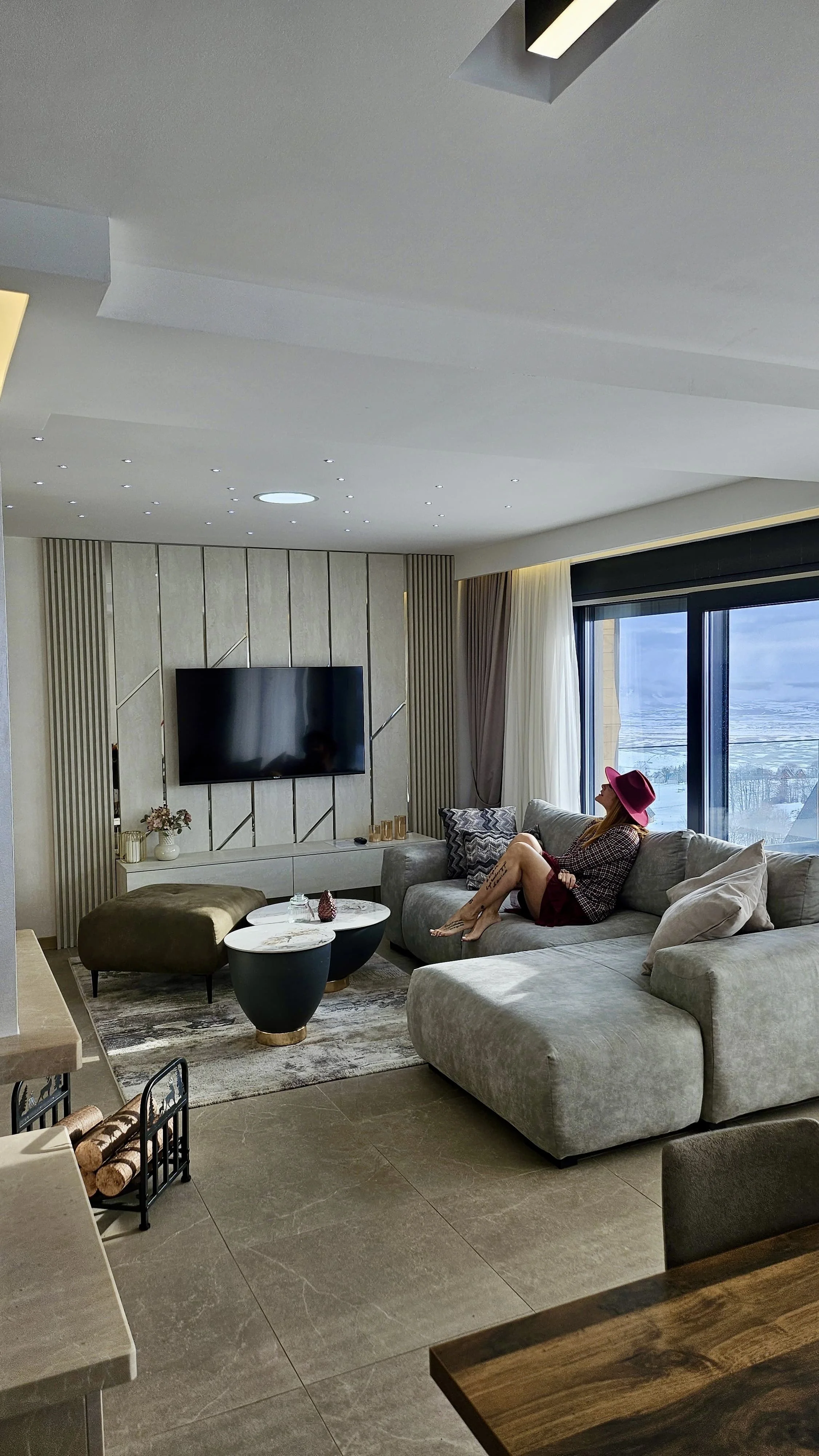 A woman wearing a red hat and patterned top sitting on a beige sofa in a modern living room with large windows, with an ocean view outside.