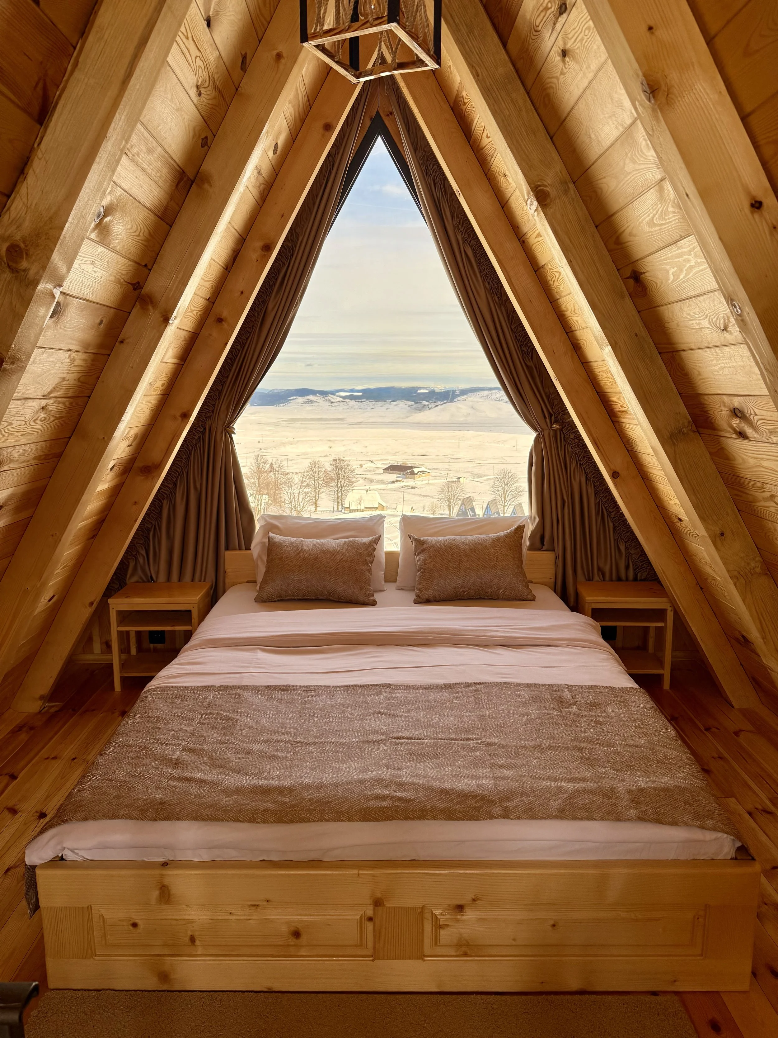 A cozy bedroom with a bed featuring white sheets, beige pillows, and a beige blanket, inside an A-frame wooden cabin. The room has a large triangular window with curtains, overlooking a snowy landscape with mountains and trees in the distance.