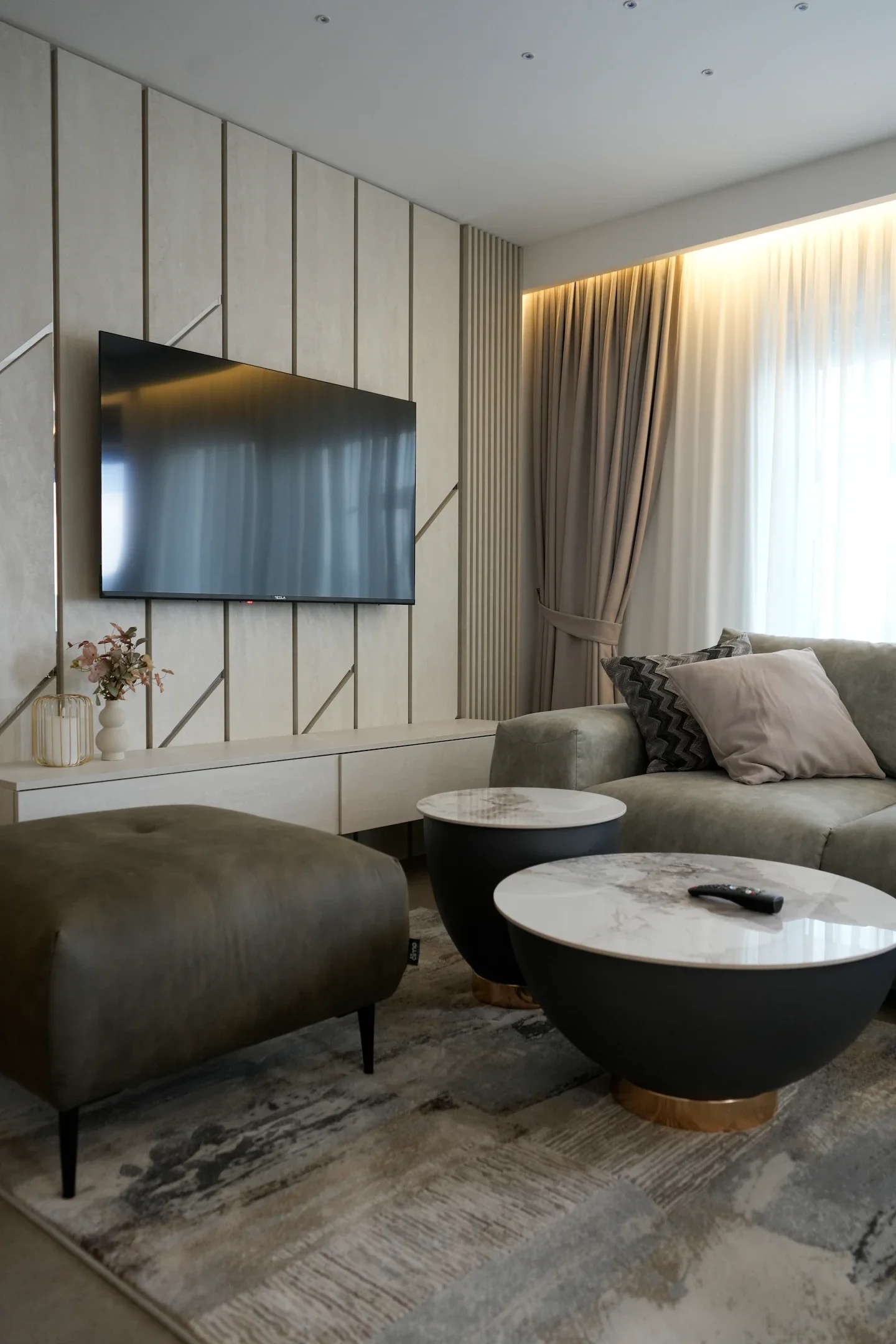 Modern living room with a mounted flat-screen TV, beige textured wall, curtains, beige sofa with cushions, marble-topped coffee tables, and a rug.