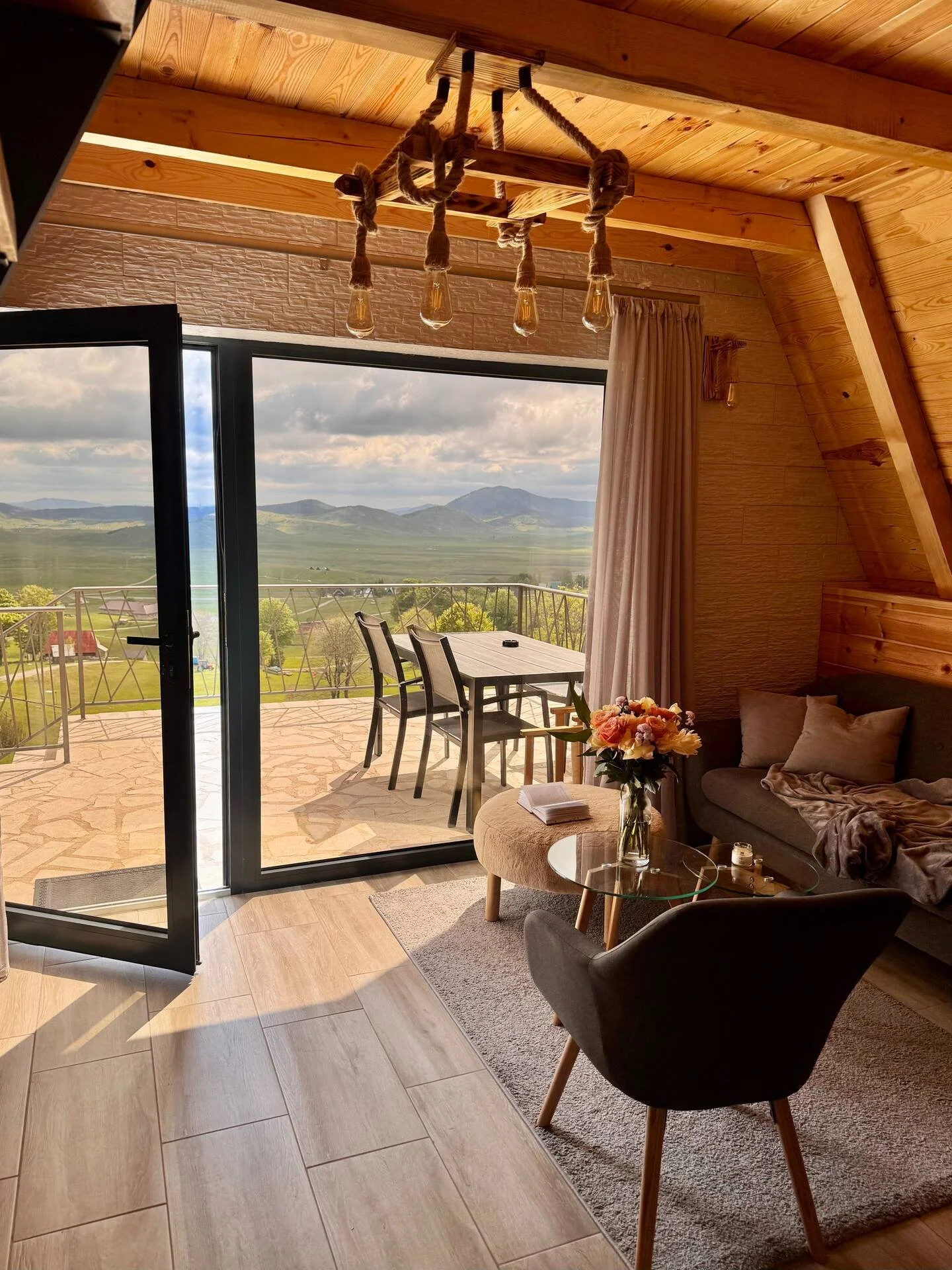 Cozy living room with wooden walls and ceiling, outdoor patio with table and chairs, mountain landscape view, glass sliding door, sofa with pillows, coffee table with flowers, and soft lighting.