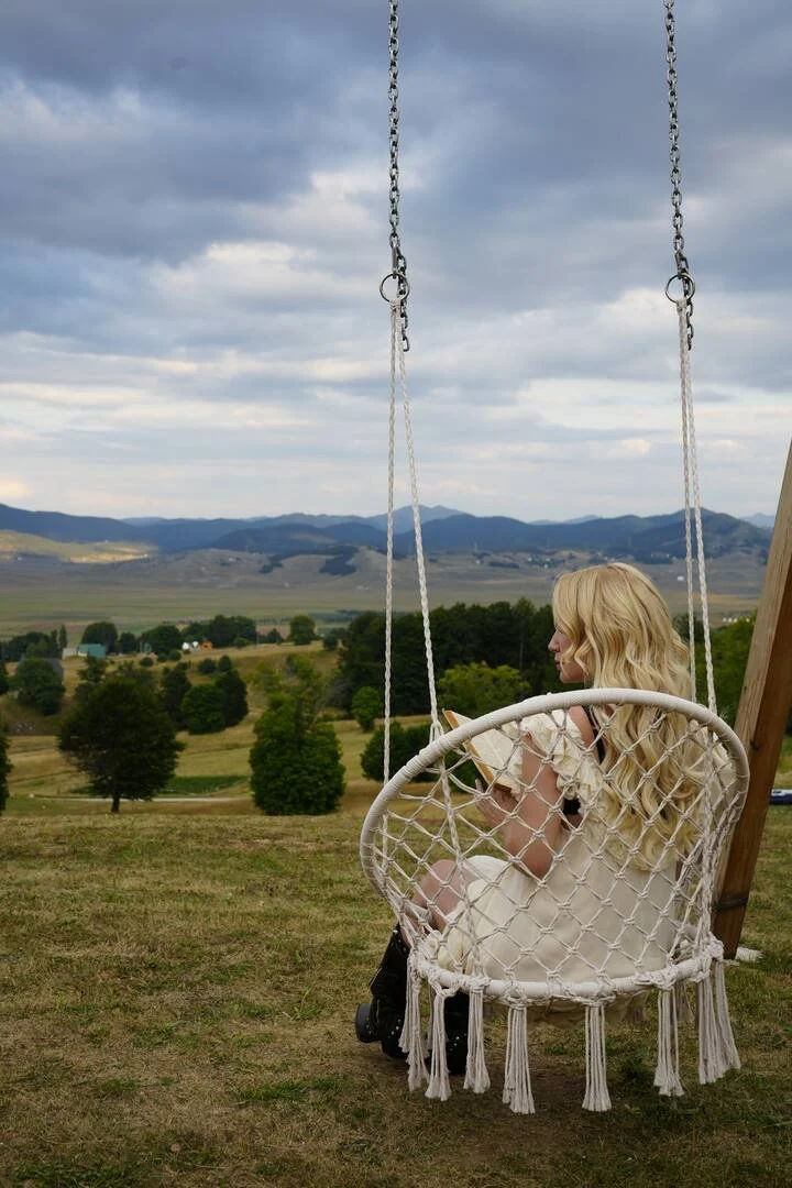 A woman with long blonde hair sits on a hanging swing chair on a grassy hill, reading a book. The swing has a woven design with tassels at the bottom, and it is set against a scenic landscape of rolling hills and mountains under a cloudy sky.
