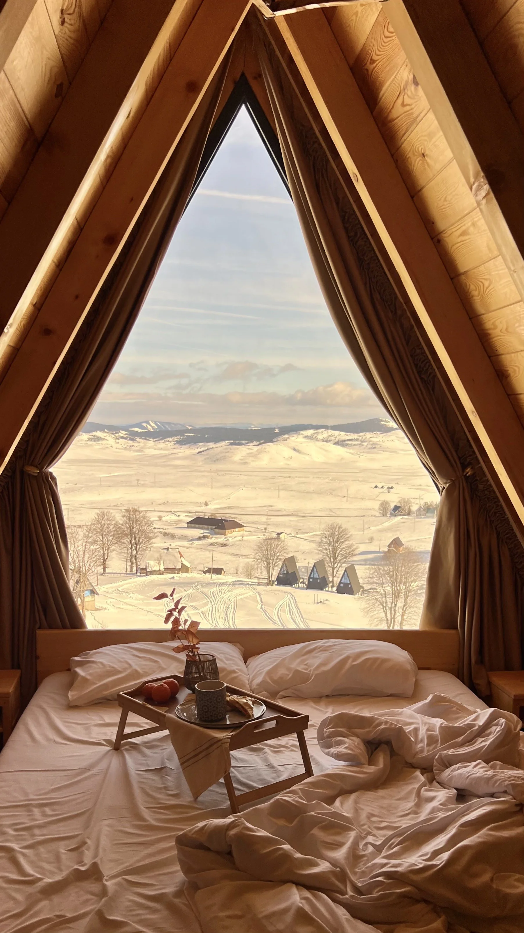 A cozy bed inside a wooden cabin with a large triangular window showing a snowy landscape with rolling hills, farm buildings, and trees.