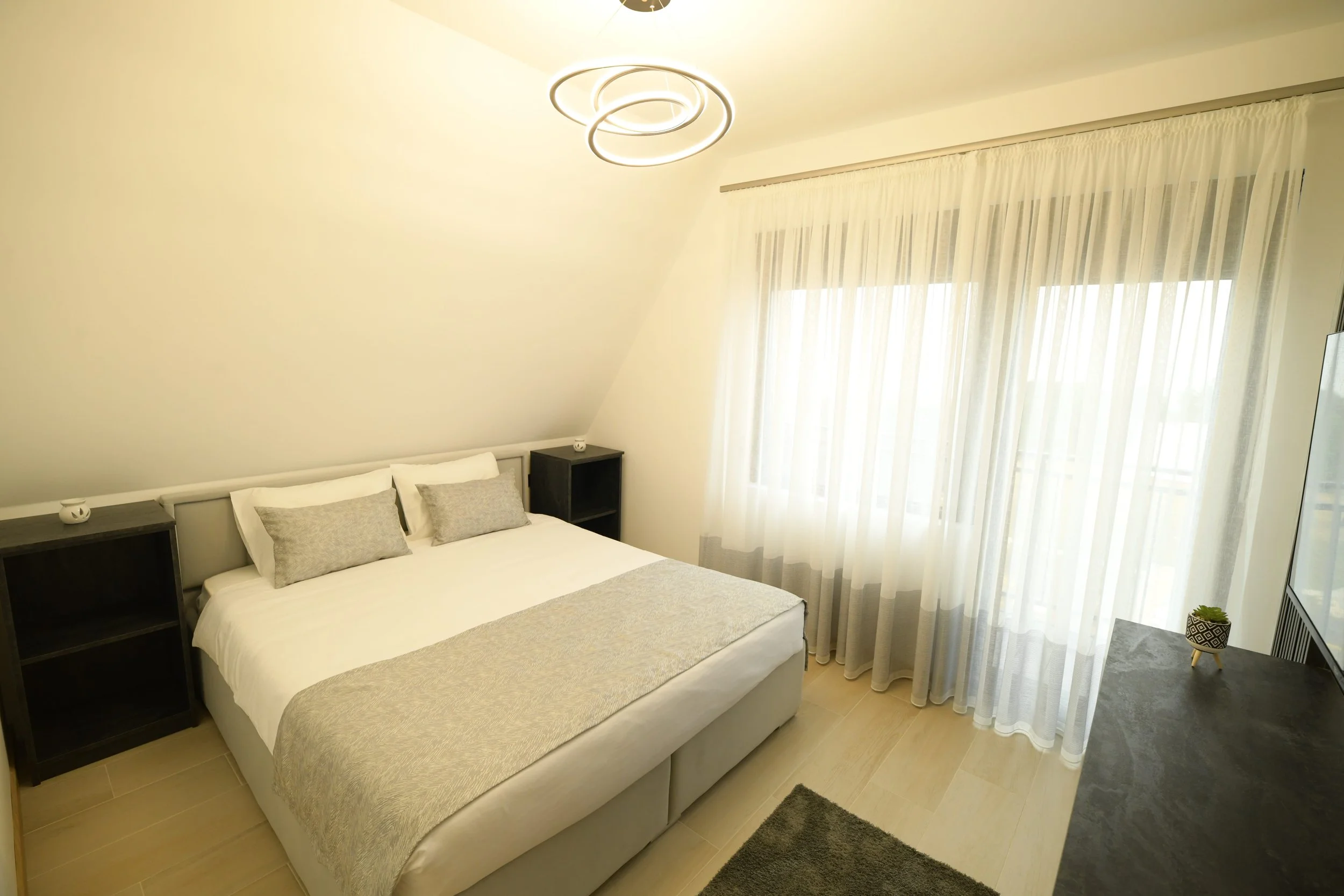 Bright bedroom with a large bed, gray and white bedding, black side tables, sheer curtains, and a modern ceiling light fixture.