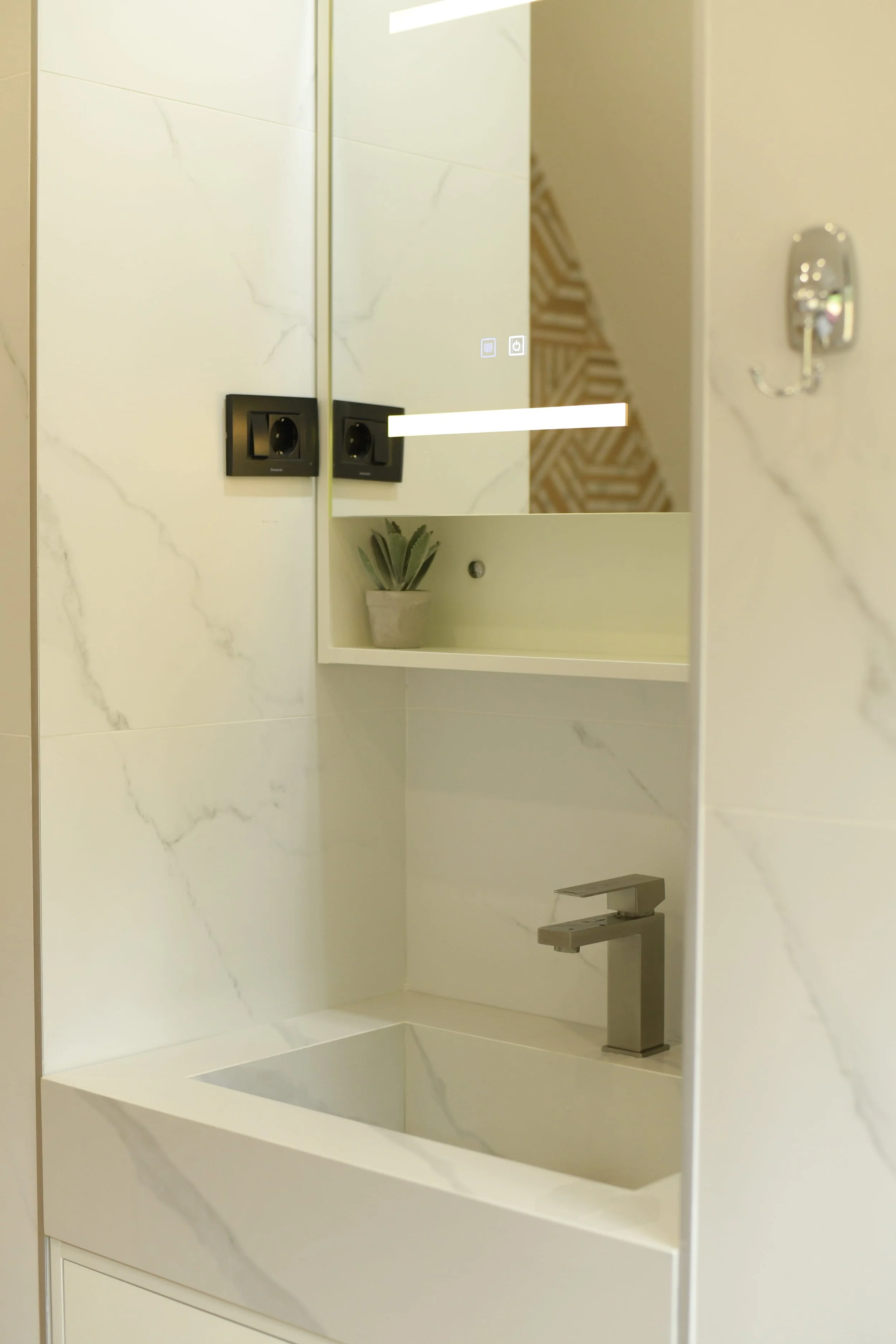 A small bathroom sink with a modern faucet, a wall-mounted mirror, a small shelf with a potted plant, and electrical outlets on a marbled wall.