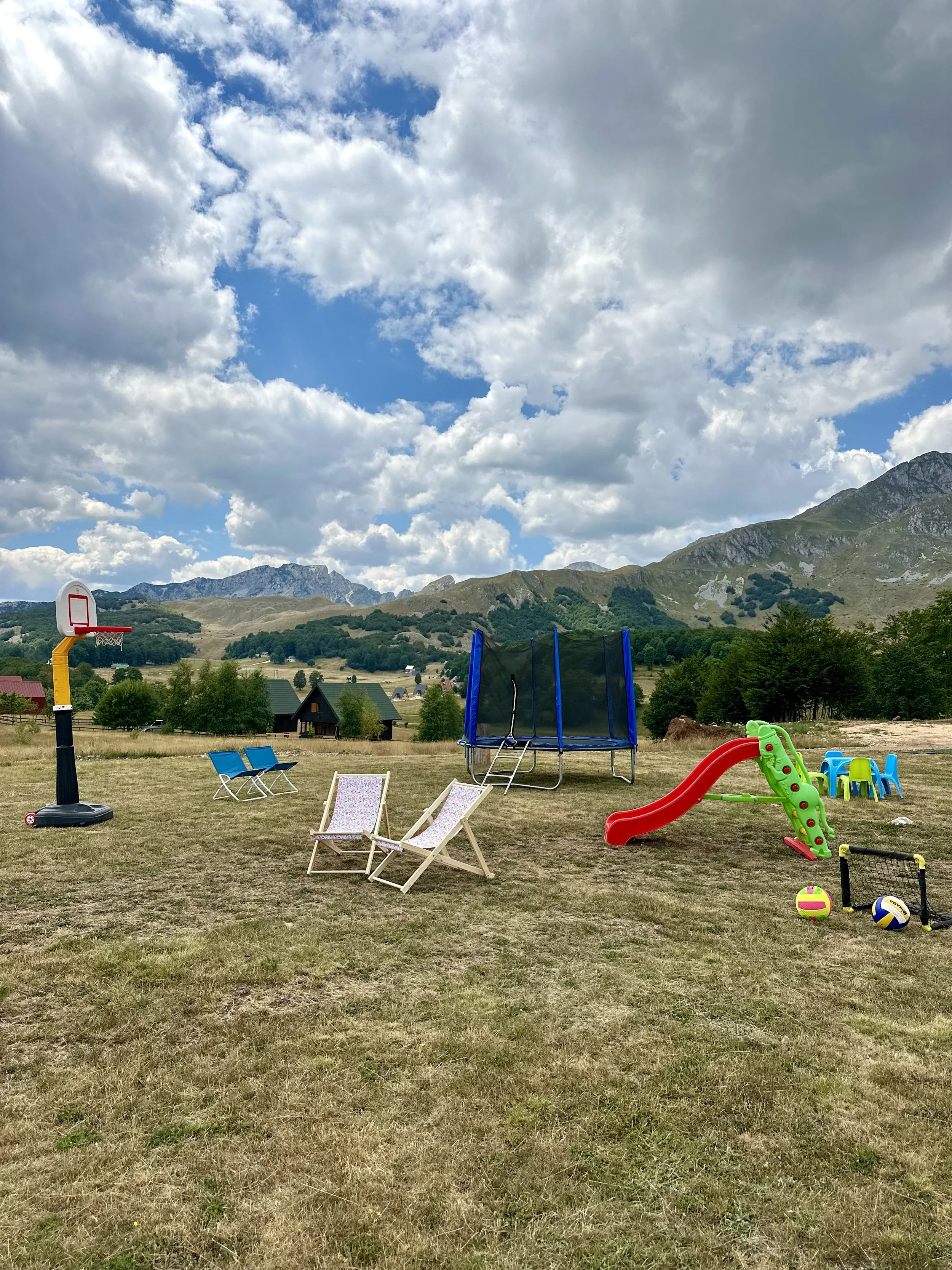 A backyard with a basketball hoop, lawn chairs, a trampoline, a slide, small tables and chairs, and various sports balls against a mountain range and cloudy sky.