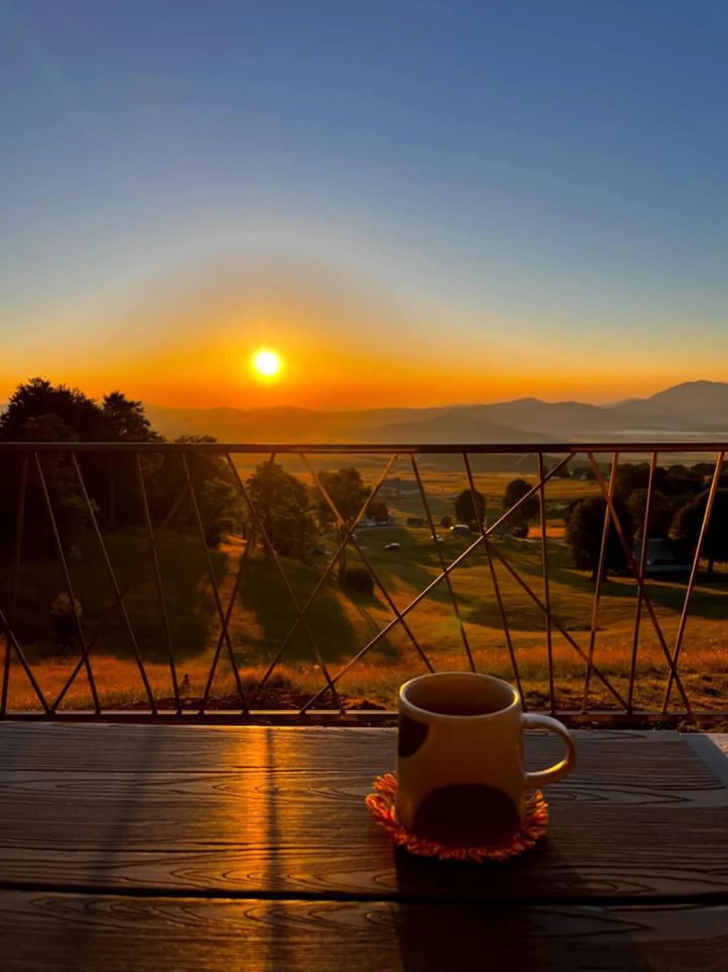 A sunrise over a scenic landscape with rolling hills, trees, and a distant mountain range. On a wooden table with a cup of coffee or tea, overlooking the view through a balcony railing.