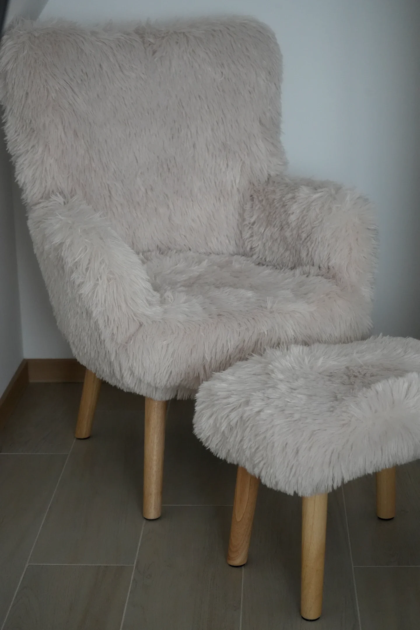 Cream-colored plush armchair with wooden legs and matching ottoman, both covered in fuzzy fabric, placed on a tiled floor against a white wall.