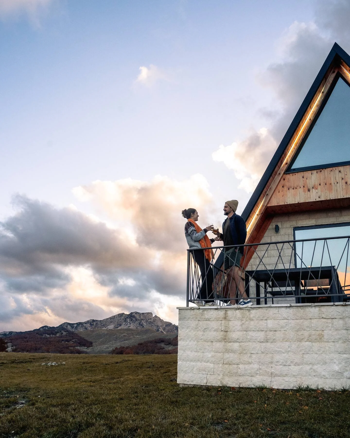 A couple standing on a balcony outside a modern house, holding drinks and talking with each other, with a mountainous landscape and cloudy sky in the background.