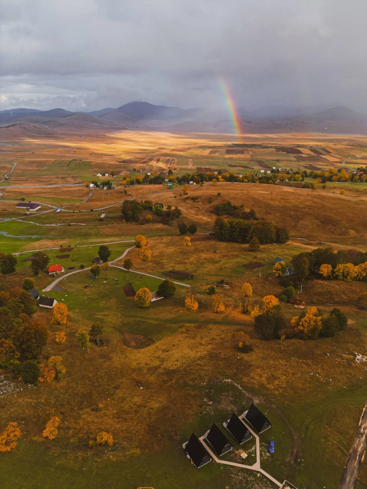 An aerial view of a rural landscape with scattered houses, trees with autumn foliage, winding roads, and a rainbow appearing over distant mountains under a cloudy sky.