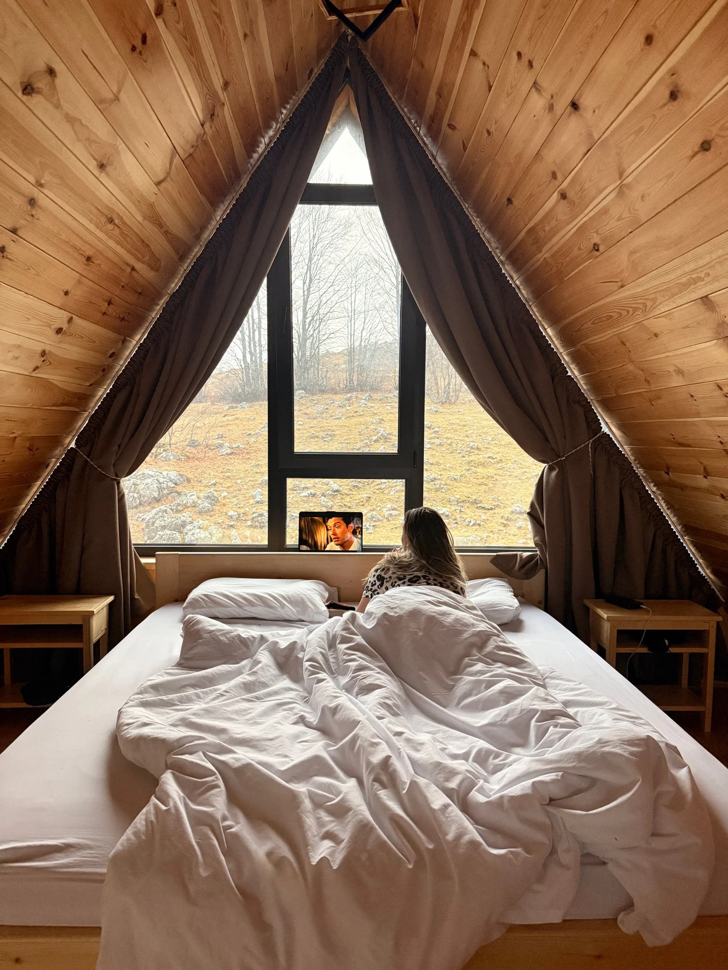 A person with long blond hair sitting on a bed with white bedding, looking out a large window in an A-frame wooden room, with a landscape of trees and rocks outside.