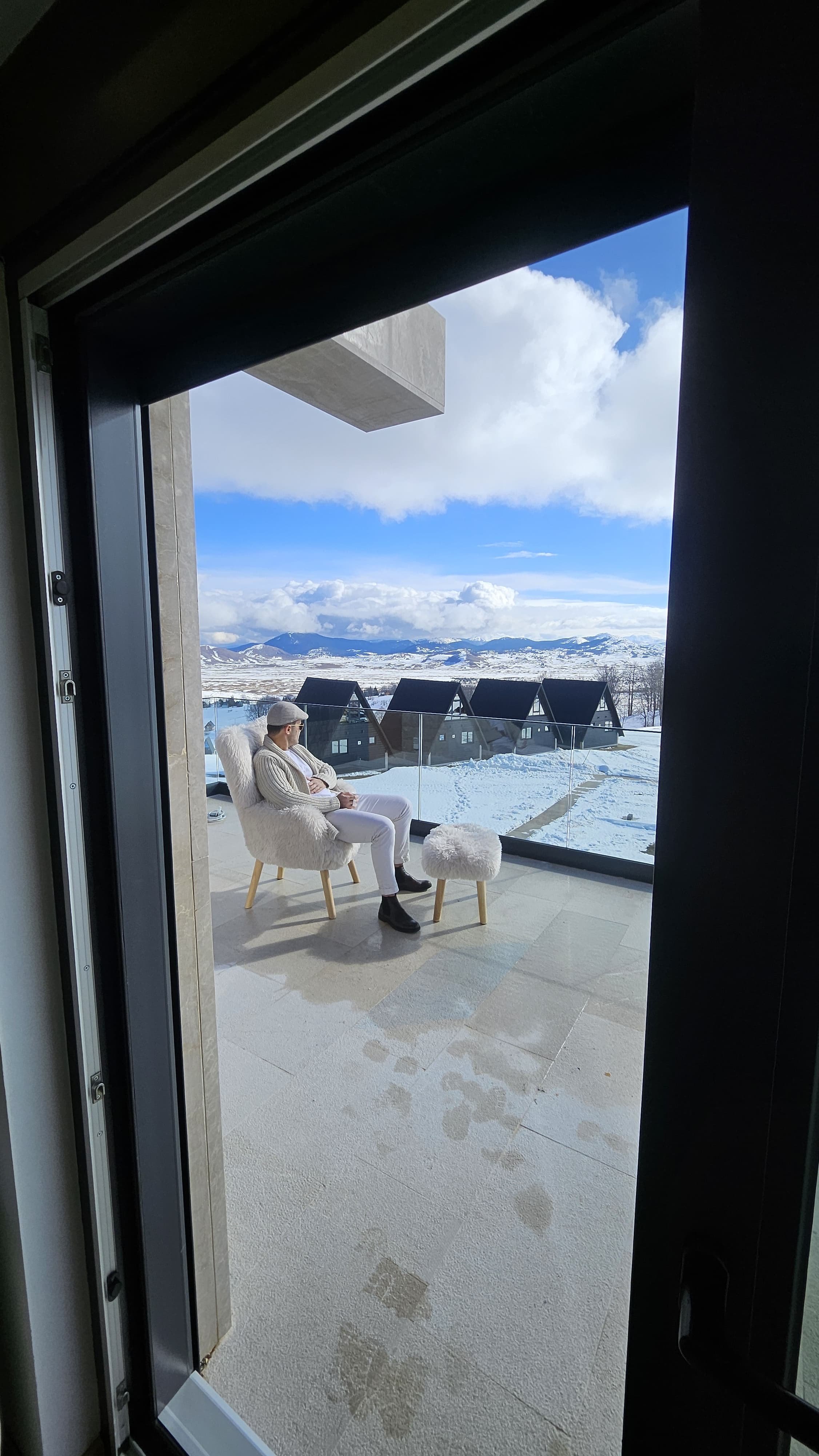 A person sitting on a fluffy white chair with a matching footstool on a balcony overlooking a snowy landscape with mountains and houses, under a partly cloudy sky.