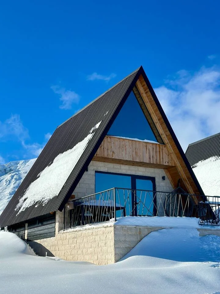 A modern A-frame house with snow on its roof, large glass doors, and a snow-covered balcony, set against a bright blue sky.