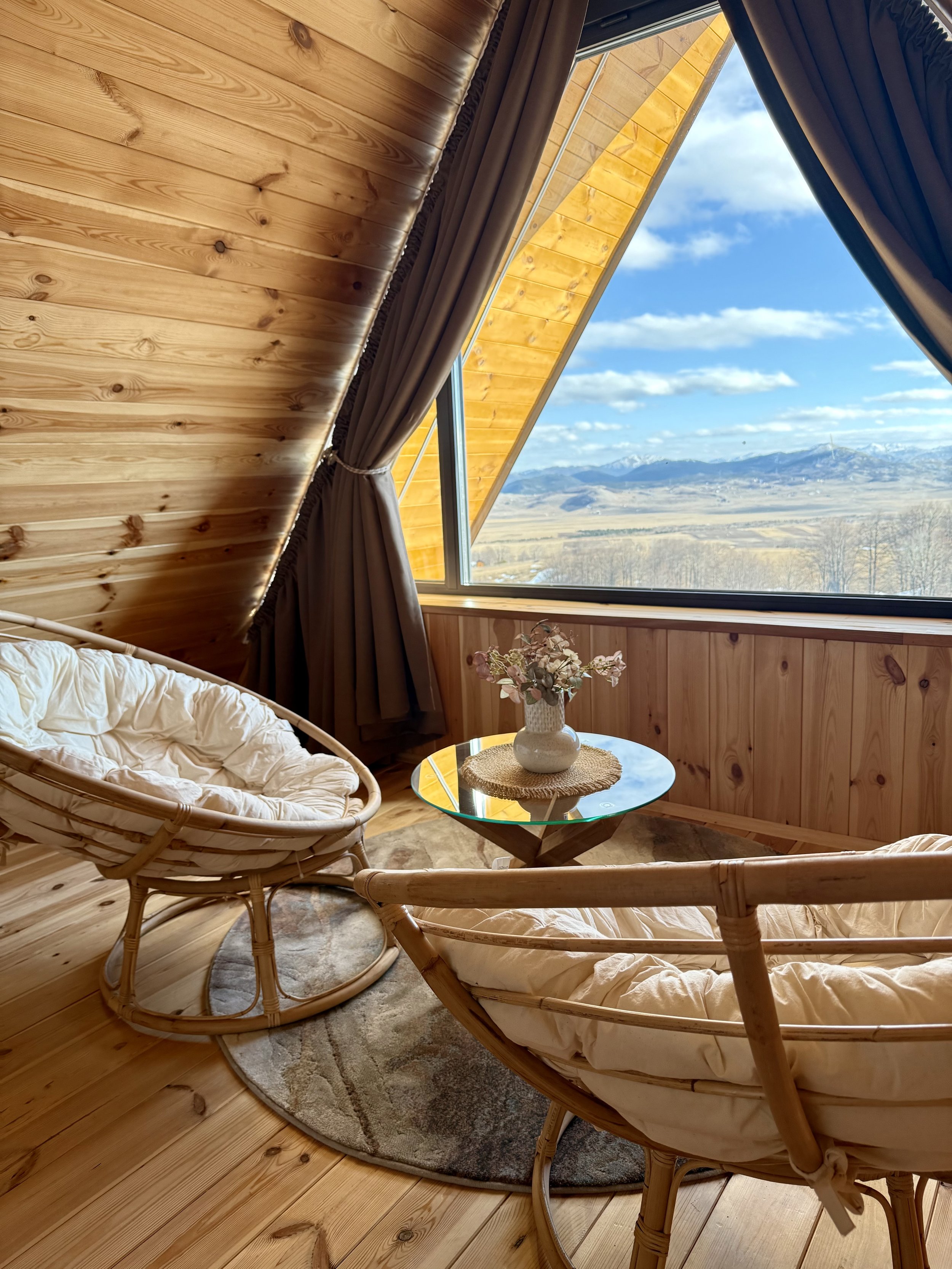 Cozy interior room with wooden walls and ceiling, featuring a large window with a view of mountains and a blue sky, a small round glass table with a flower vase, and two rattan chairs with cushions.