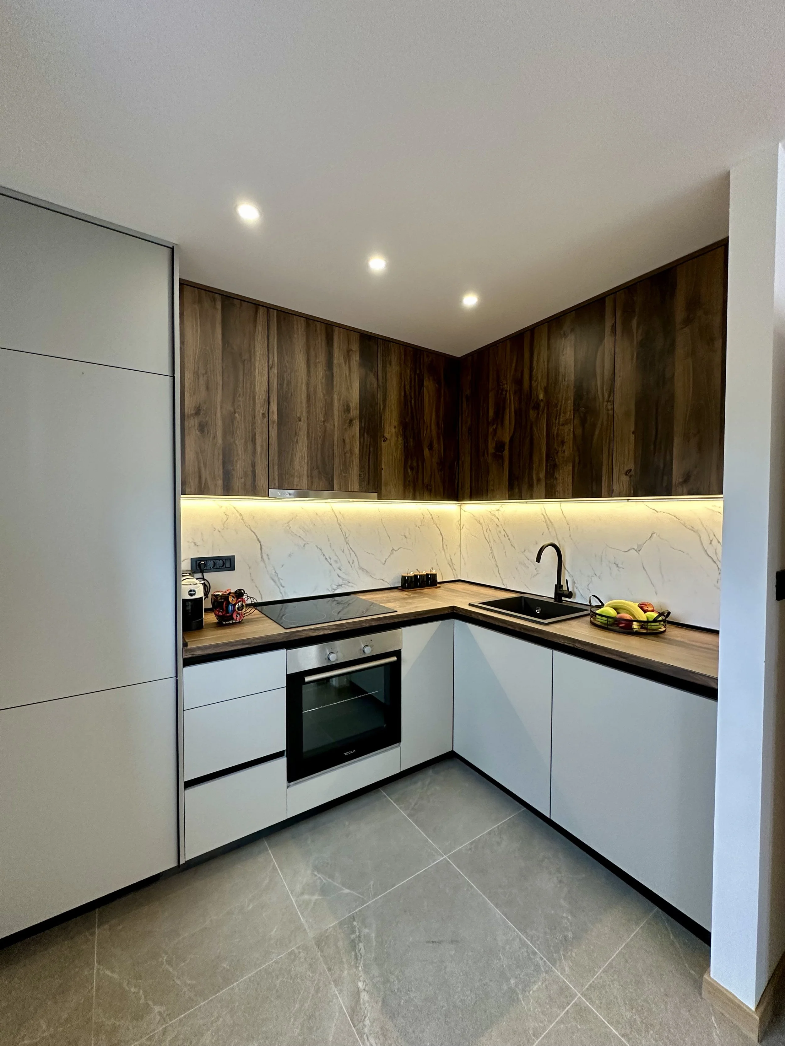 Modern kitchen with wood and white cabinets, marble backsplash, black faucet, stovetop, built-in oven, and a bowl of fruit on the countertop.