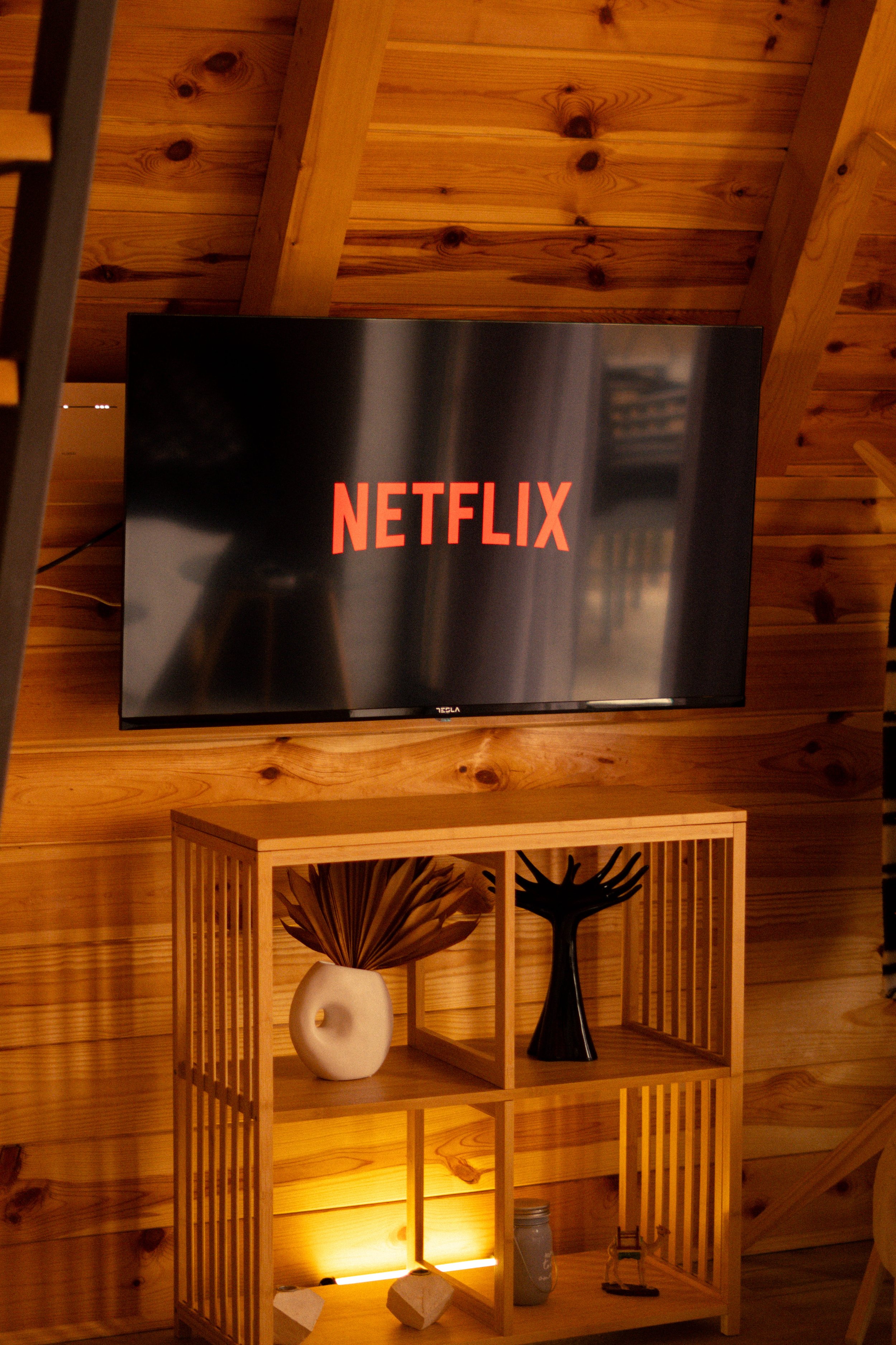 A wall-mounted flat-screen TV displaying the Netflix logo, situated above a wooden shelving unit with decorative vases and items inside, against a wooden-paneled wall.
