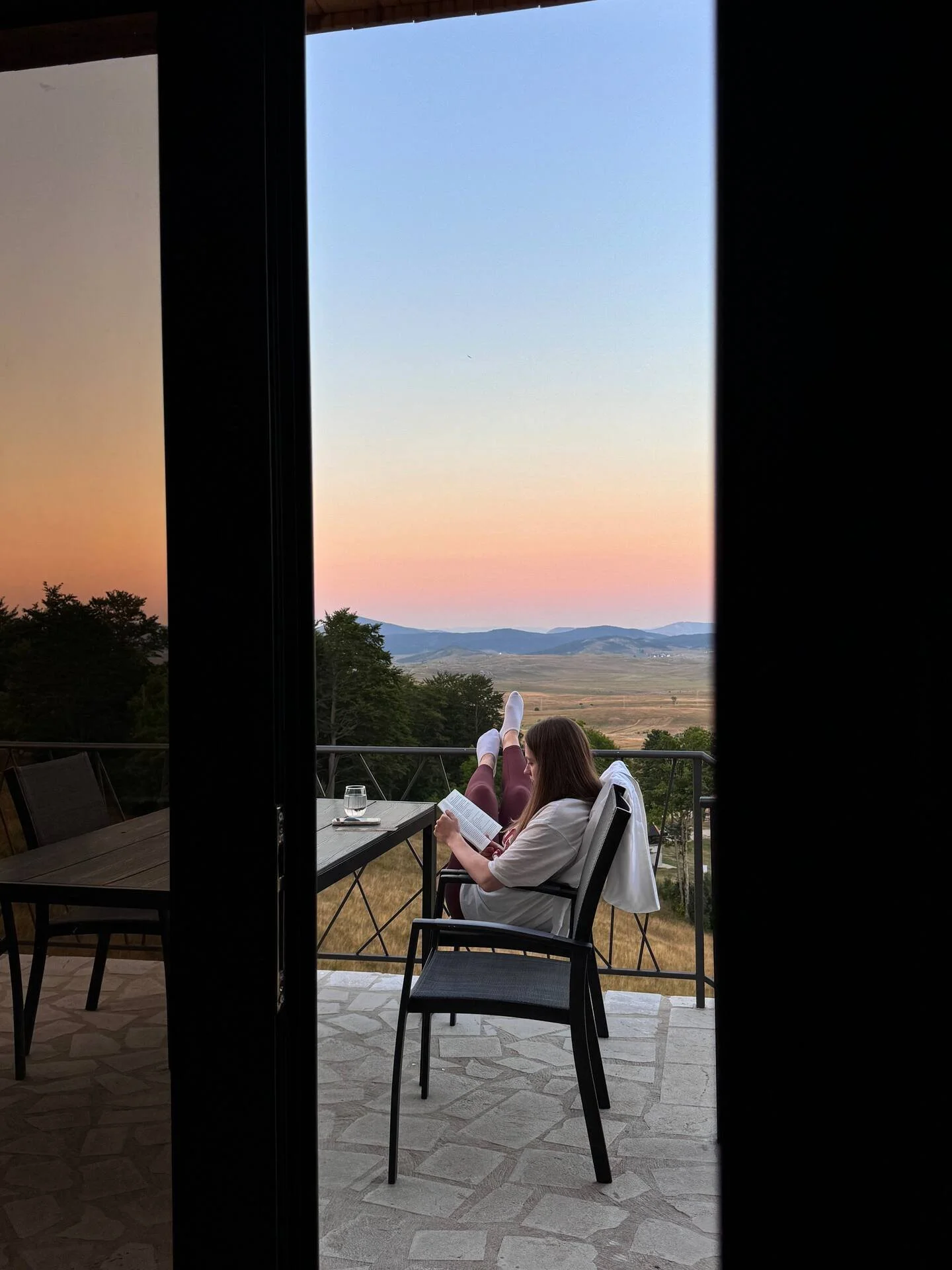 A woman sitting on a balcony at sunset, reading a book with a glass of water next to her.