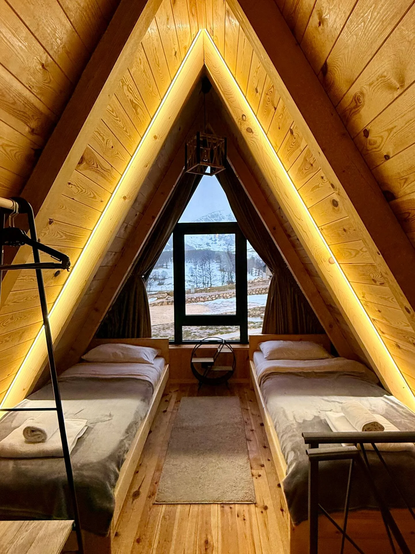 Inside an A-frame cabin with two beds, a window showing a snowy mountain landscape, and warm yellow lighting along the wooden walls.