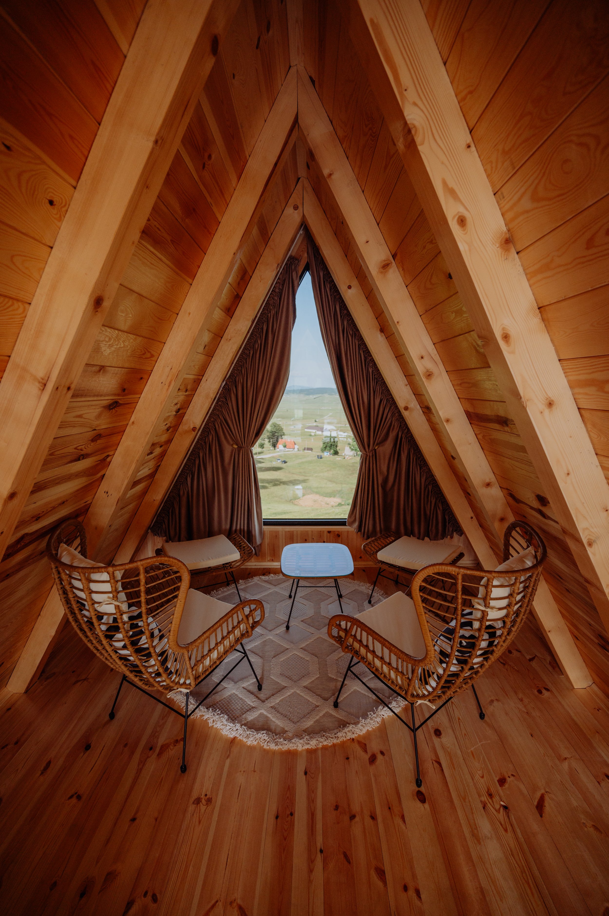 Interior of a cozy wooden A-frame cabin with a triangular window, four wicker chairs around a small table, and a view of the countryside outside.
