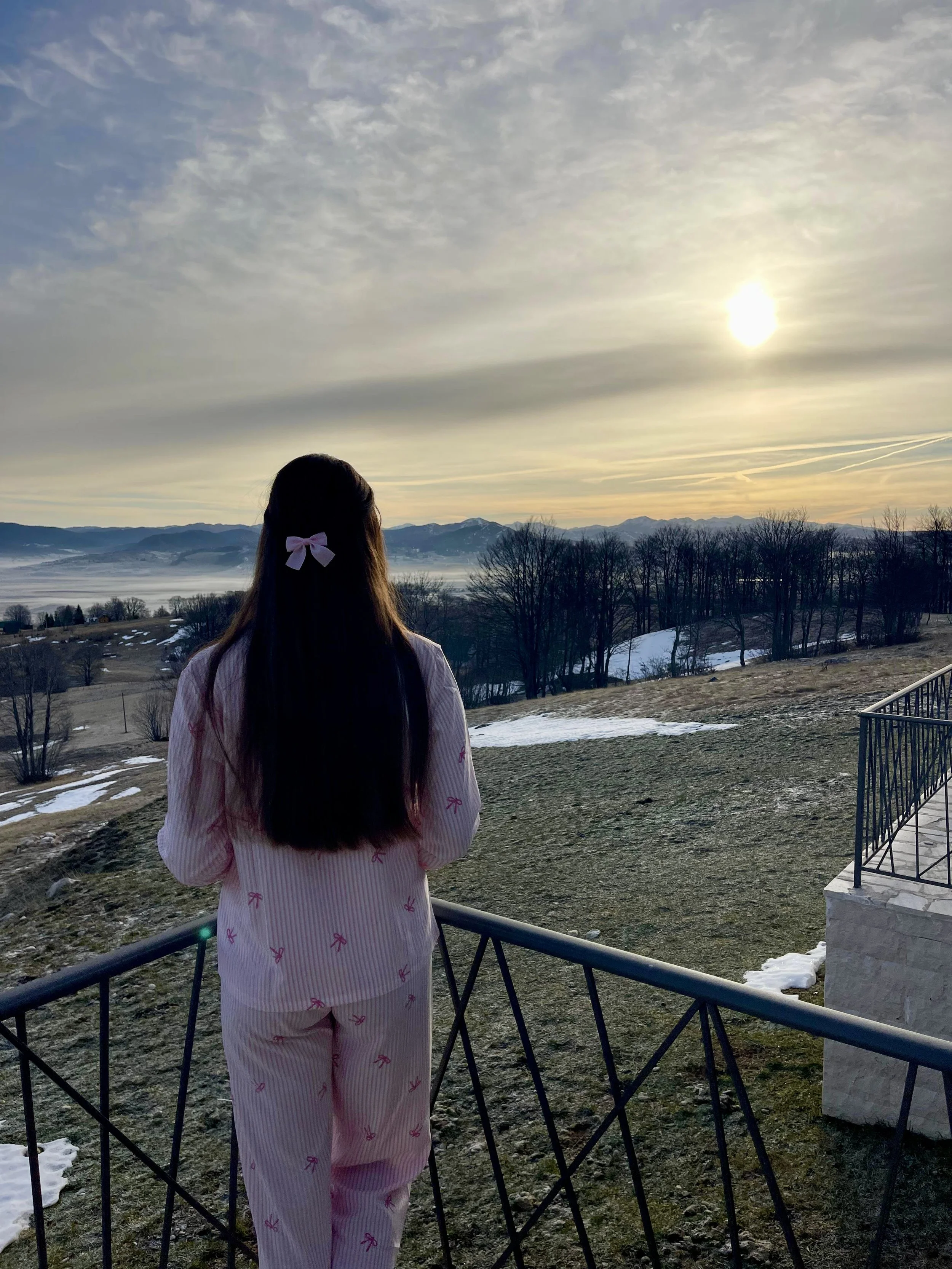 A girl with long dark hair wearing pink pajamas with small pink bows, standing on a balcony looking at a scenic winter landscape with mountains, bare trees, patches of snow, and a cloudy sky at sunset.