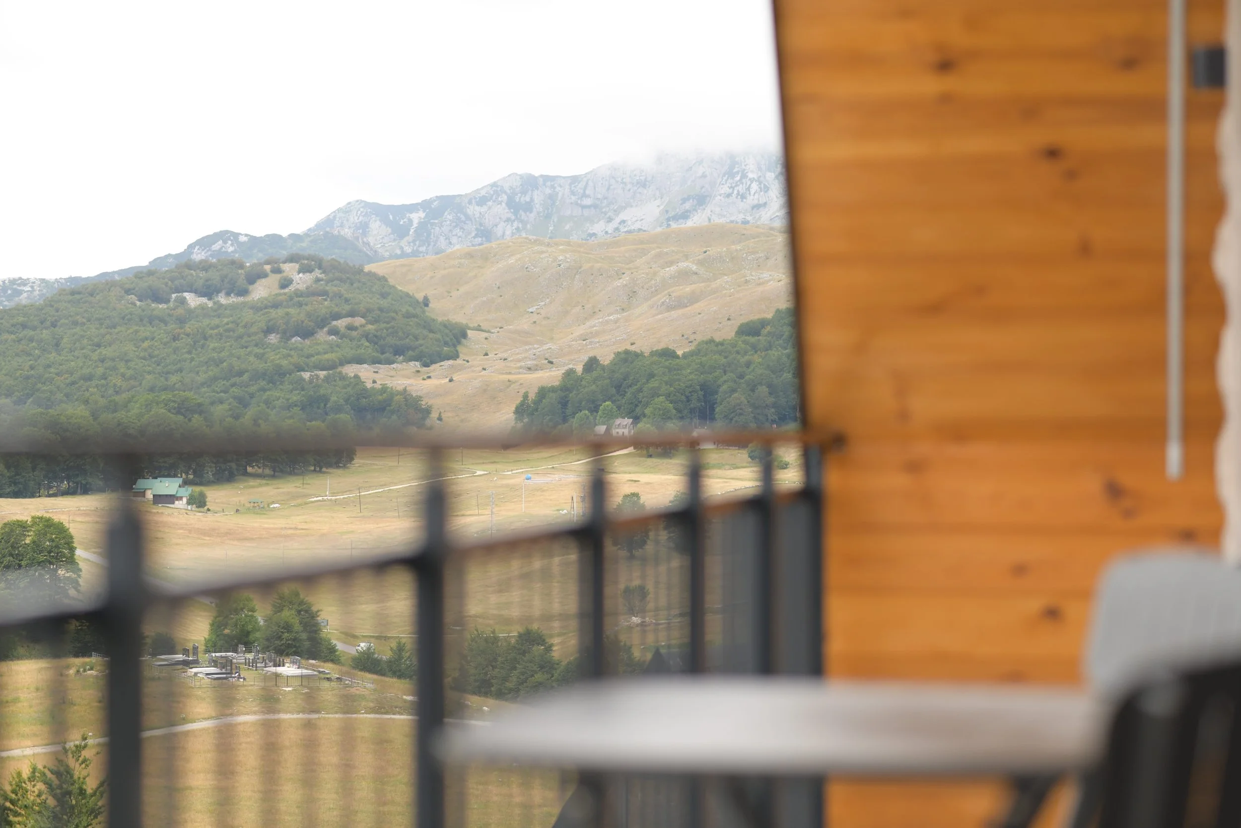 A view from a balcony showing a landscape with rolling hills, trees, and mountains in the distance, partially framed by a wooden wall on the right and a metal railing in the foreground.