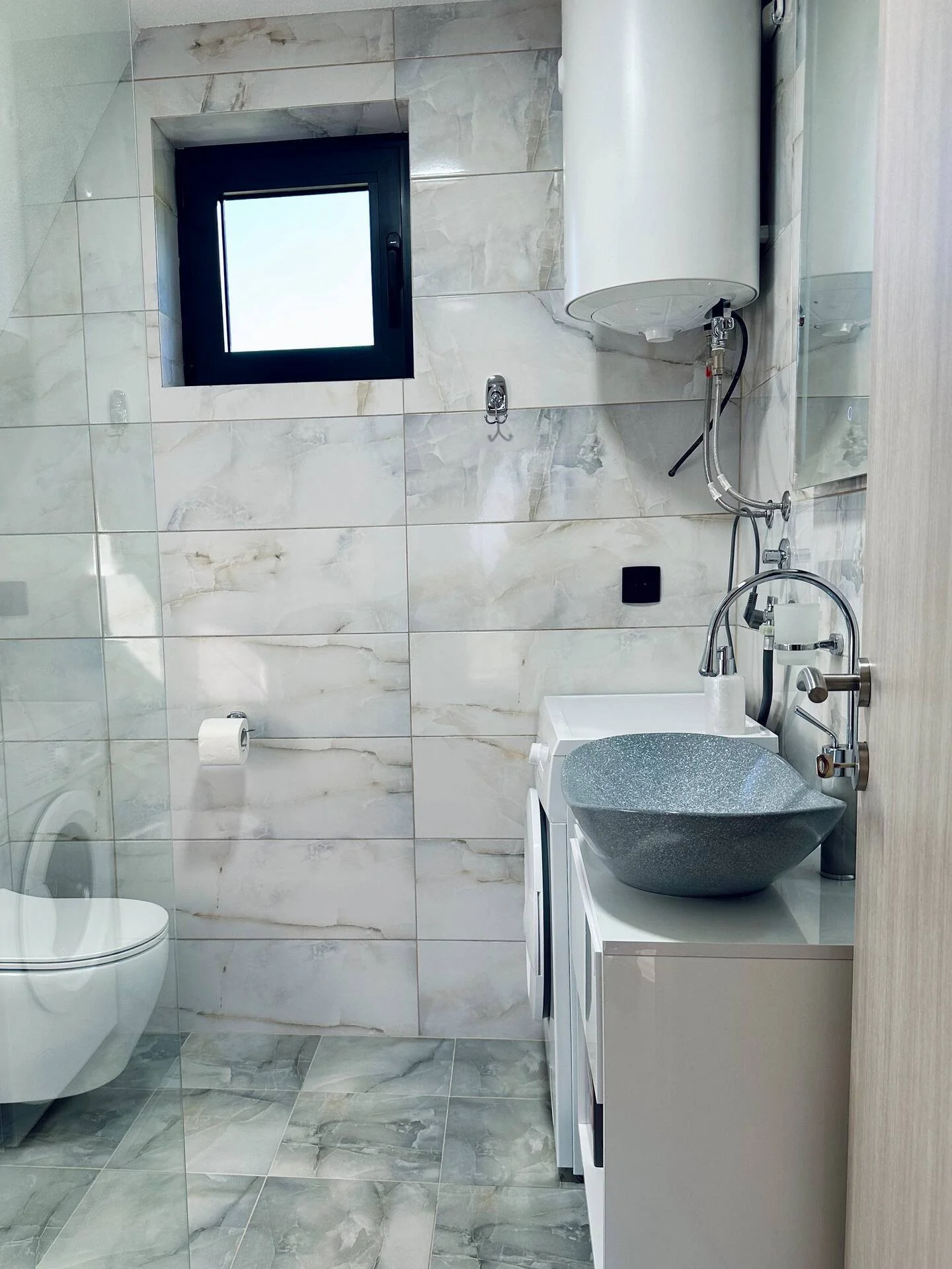 A small, modern bathroom with a toilet, a washing machine, a stone sink, a water heater, a small window, and a mirror.