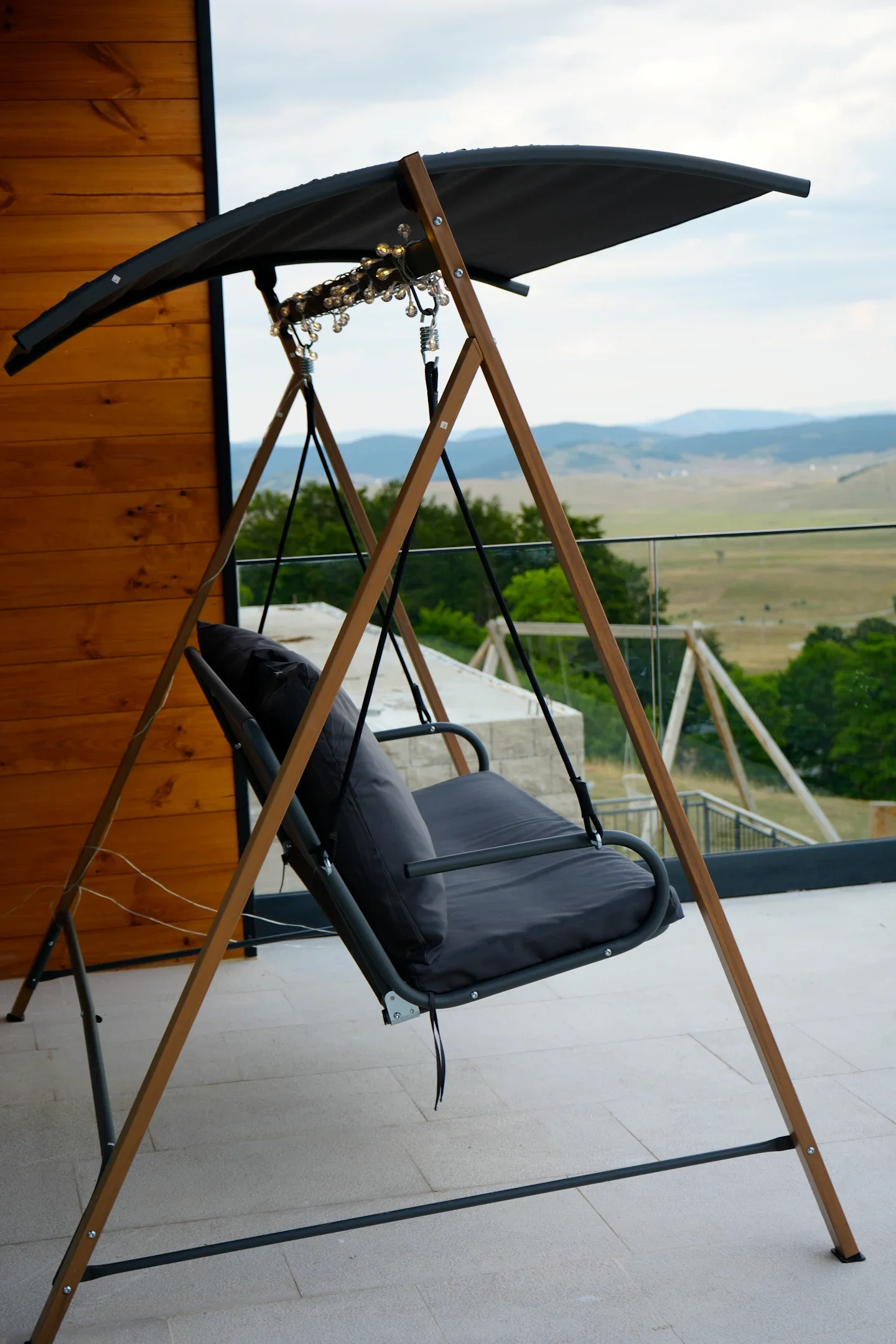A black outdoor swing with a canopy, hanging from a wooden frame, on a patio overlooking valleys and mountains in the distance.