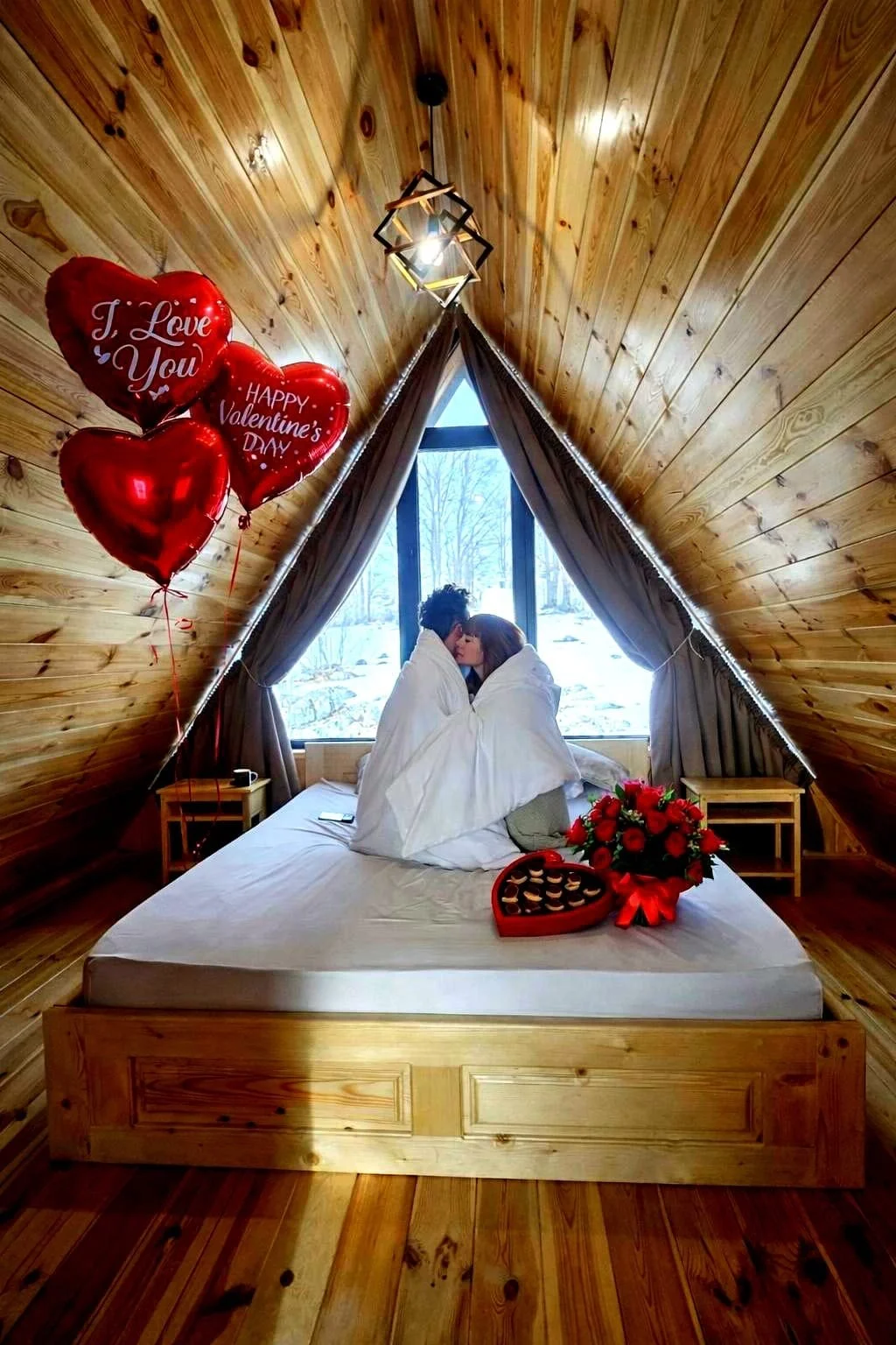 A couple wrapped in a duvet on a bed in an attic room with wood-paneled walls, a window with snowy outdoors view, red heart-shaped balloons, and a bouquet of red roses.