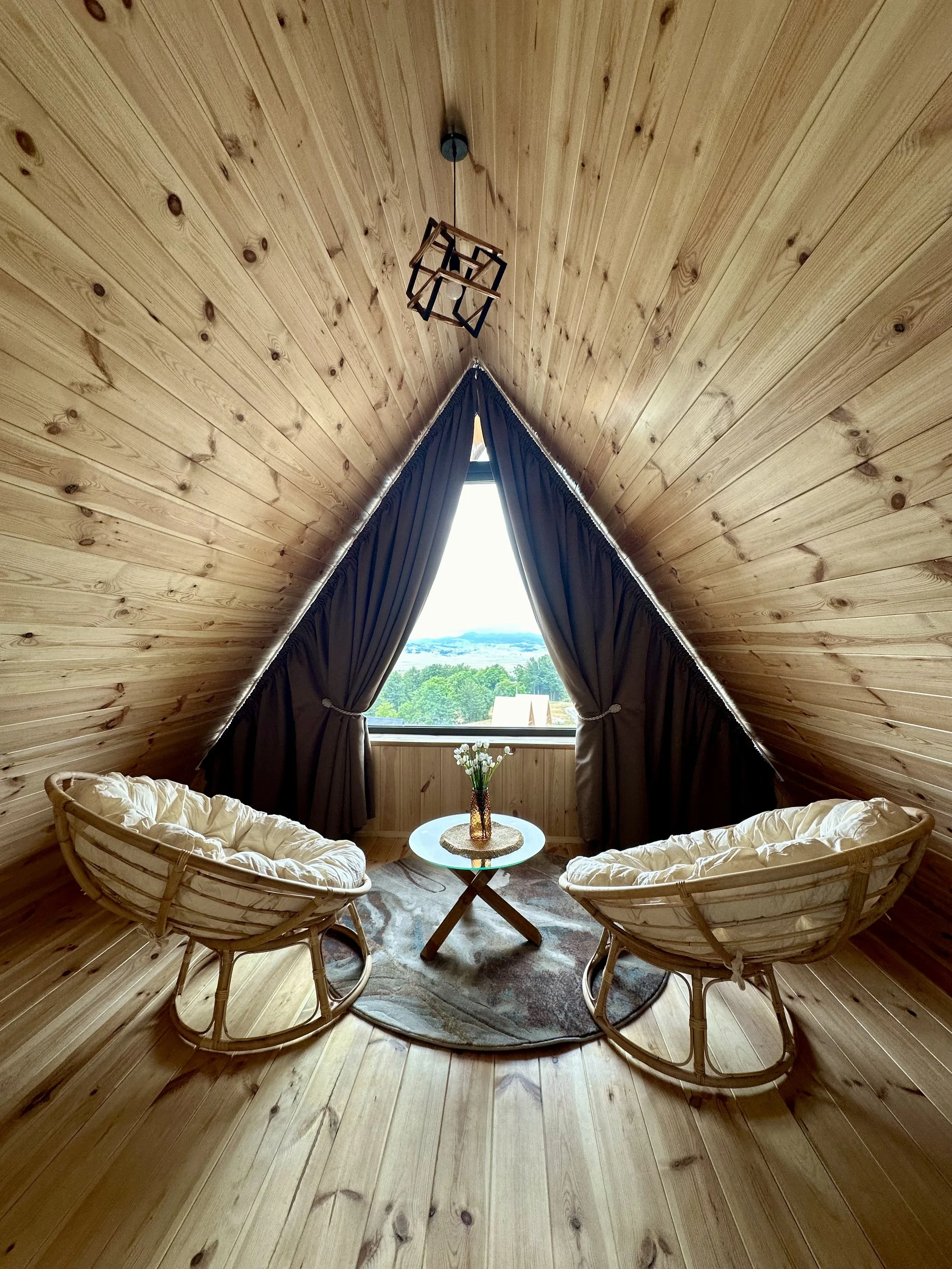 Cozy attic room with wooden walls and ceiling, two cushioned rattan chairs facing each other, a small round table with a vase of white flowers, and a large window with dark curtains, offering a view of greenery and distant landscape.