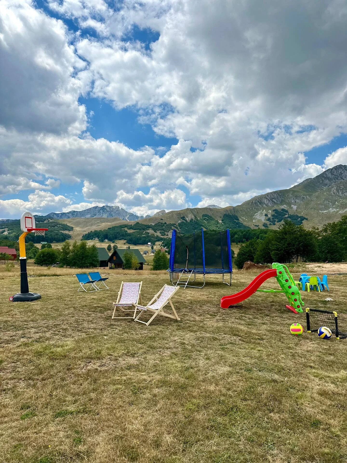 Children's outdoor play area with small slide, trampoline, basketball hoop, lounge chairs, plastic table and chairs, and sports balls on a grassy field with mountains and a partly cloudy sky in the background.