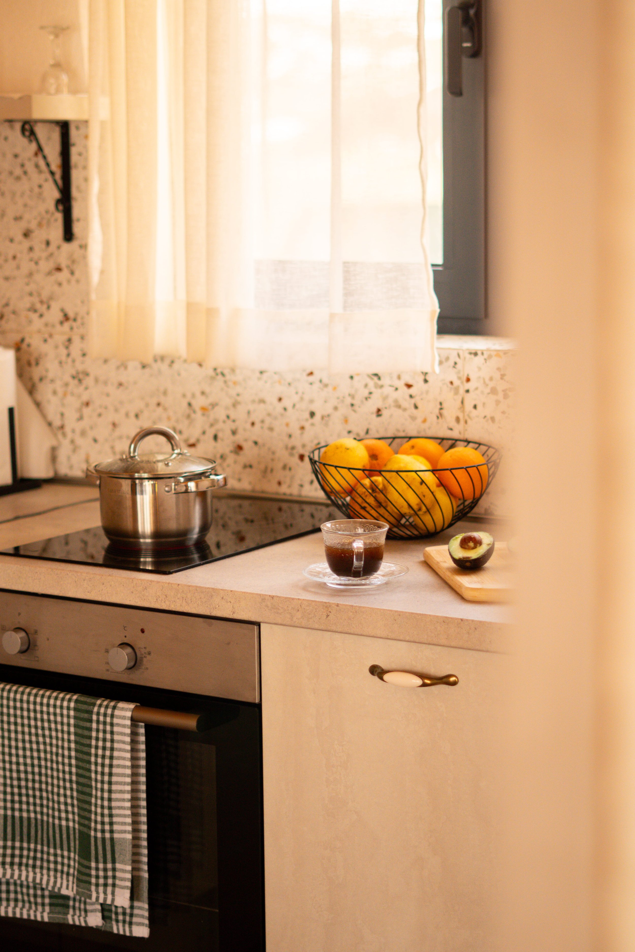 A kitchen countertop with a metal pot on a stovetop, a bowl of mixed fruits, a cup of coffee, and a sliced avocado, with a window and curtains in the background.