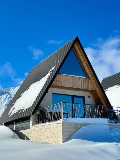A modern A-frame cabin with a balcony, snowy landscape, and mountain backdrop under a clear blue sky.