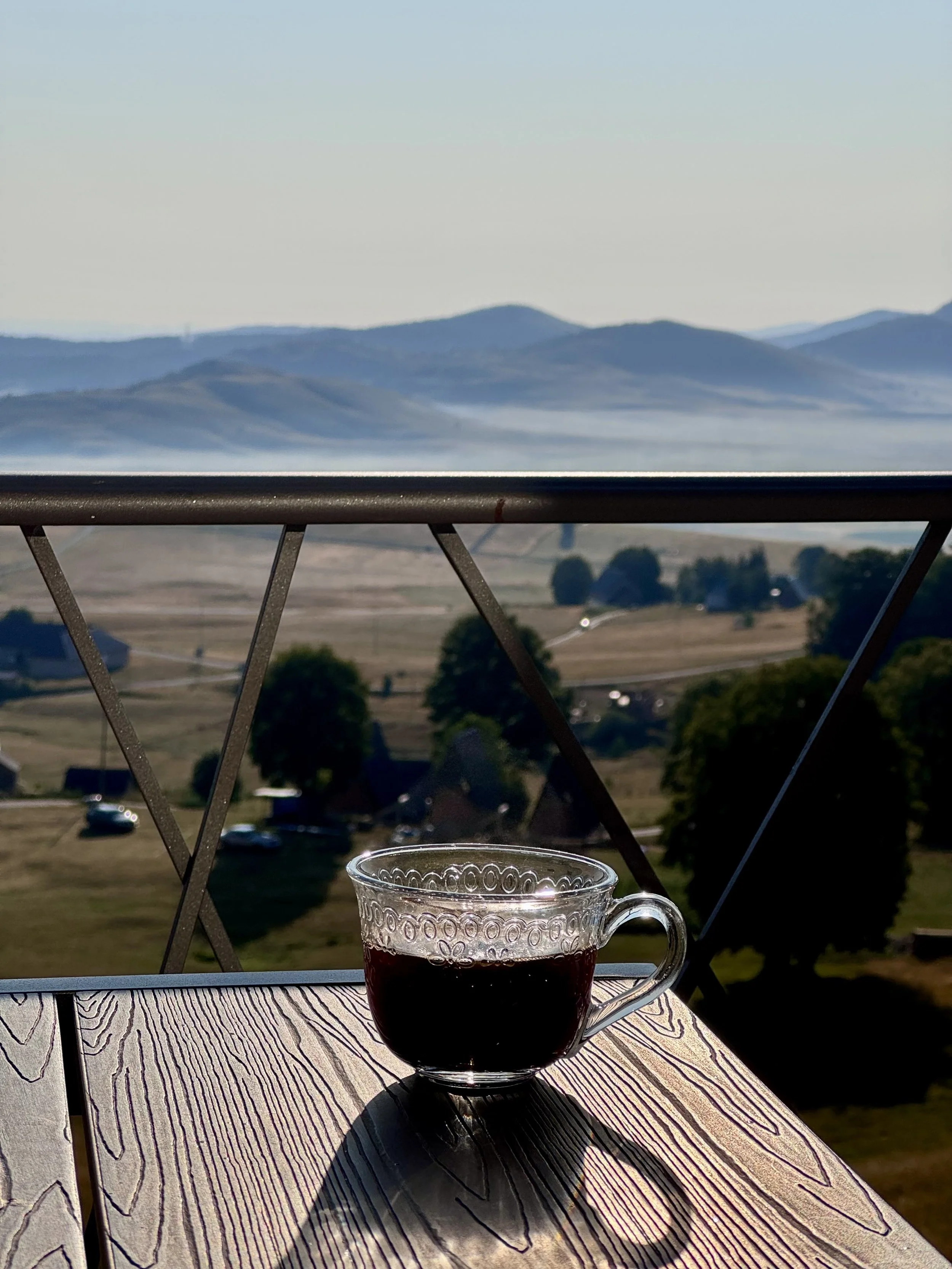 A glass cup of dark coffee on a wooden table with a shadow, overlooking a rural landscape with trees, houses, and distant mountains under a clear sky.