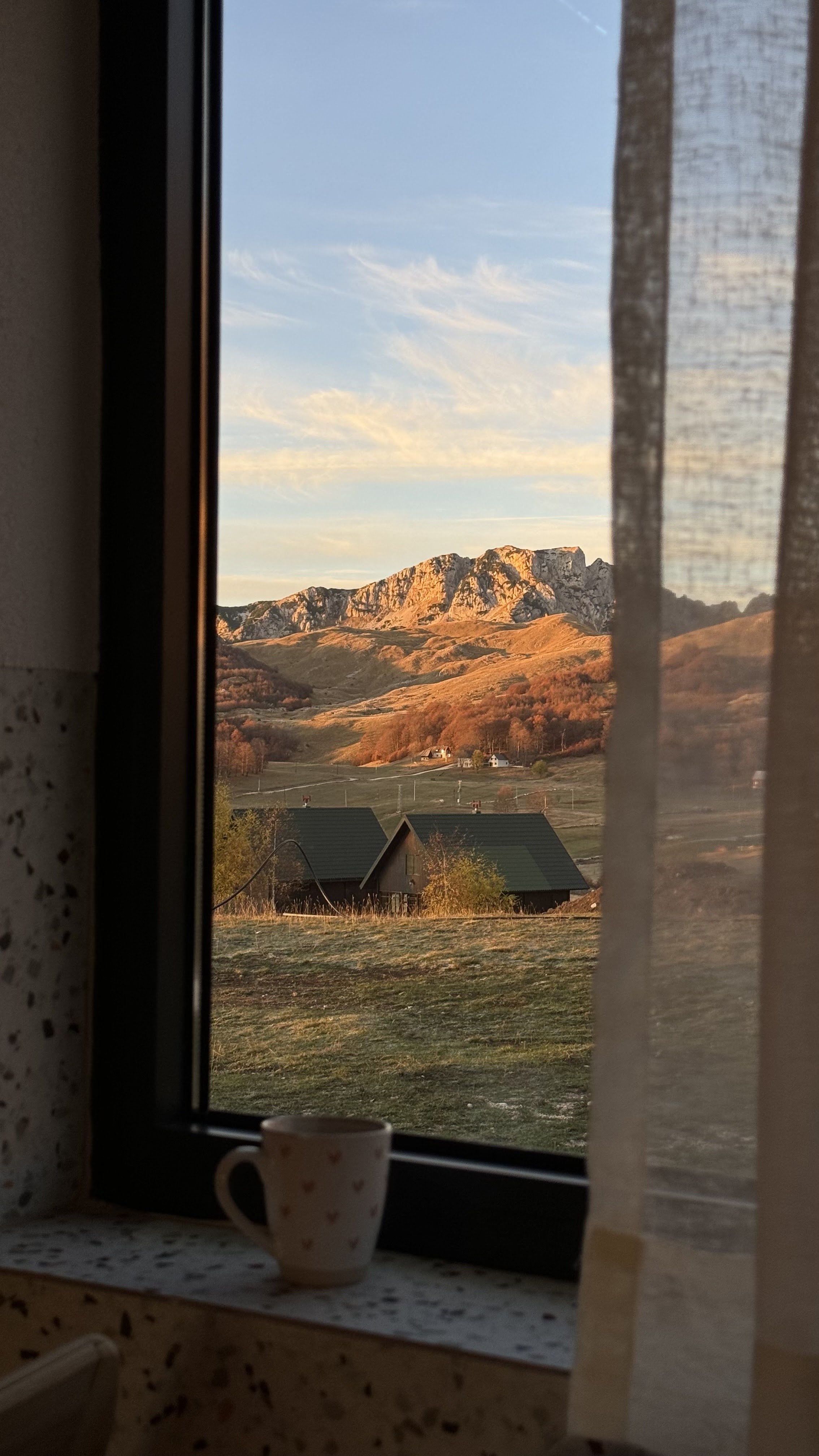 A view of mountains with rocky peaks and rolling hills, seen through a window with lace curtains, during sunset with a small house with a dark roof in the foreground, a cup on the windowsill, and a grassy yard.