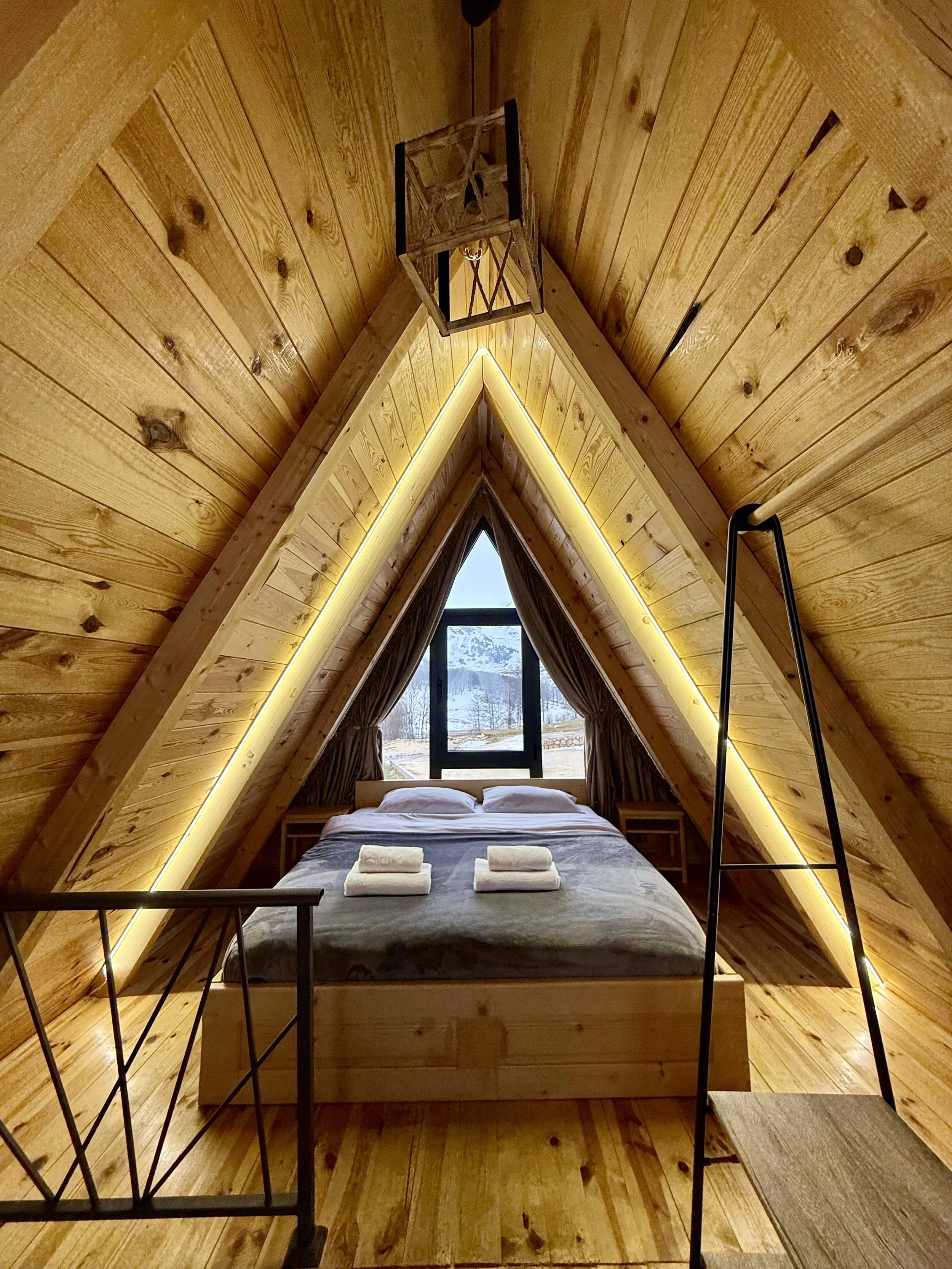 A cozy A-frame bedroom with wooden paneling, a bed with white pillows and towels, large window showing snowy mountains, and warm lighting.