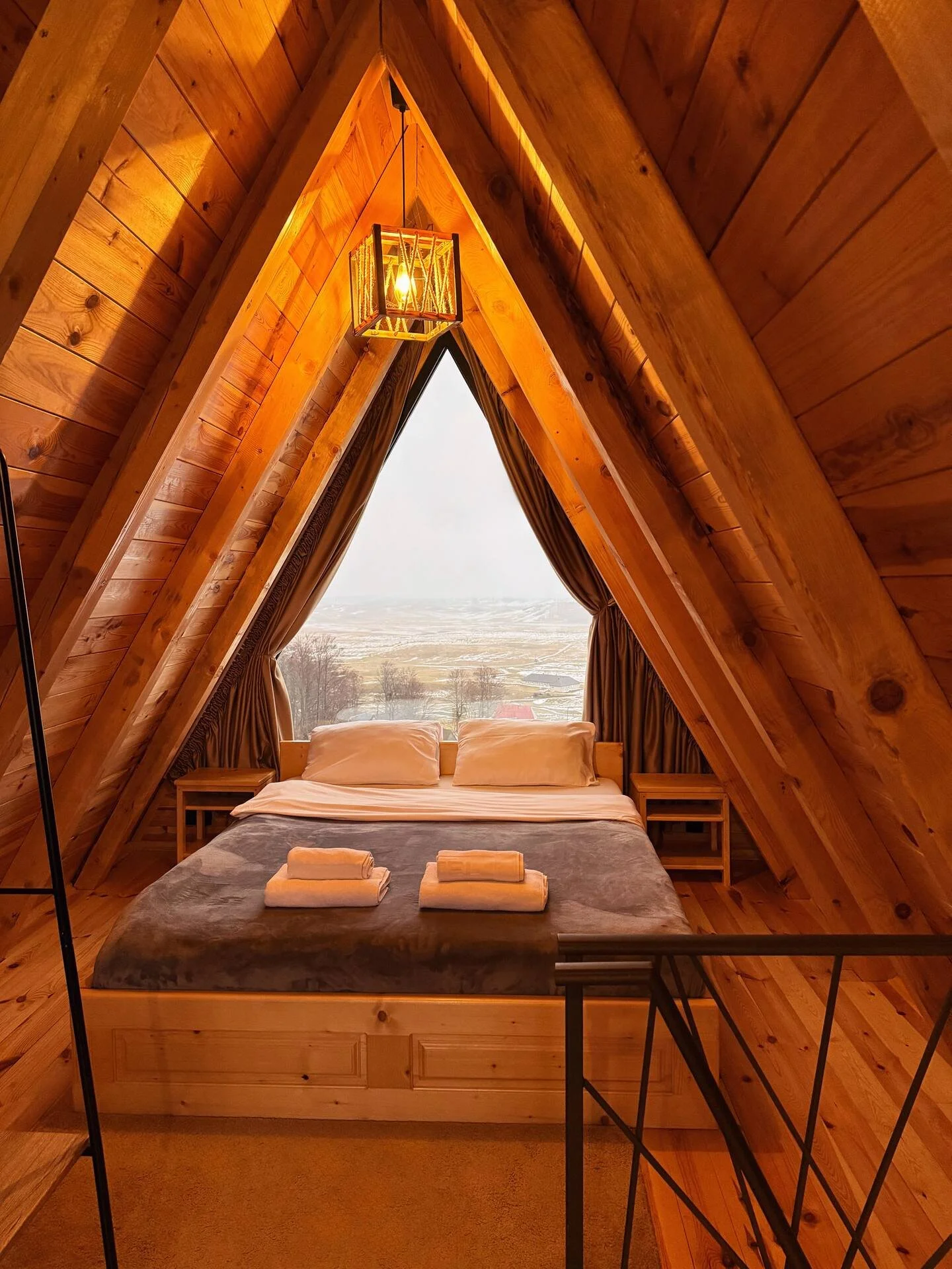 A cozy A-frame cabin bedroom with wooden walls and ceiling, a bed with white pillows and folded towels, large window with curtains revealing a snowy landscape outside, and a hanging lantern light fixture.