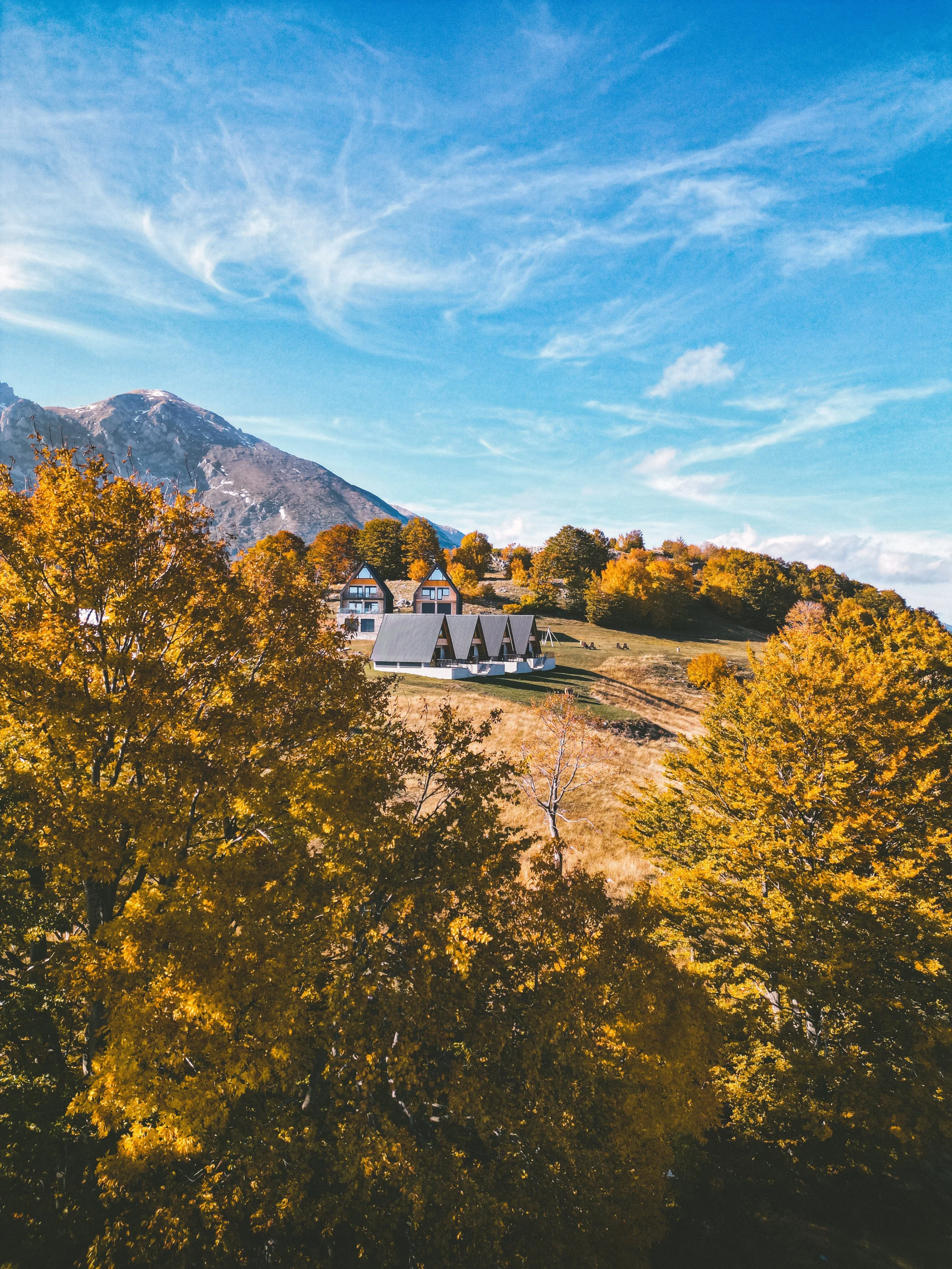 A mountainous landscape with houses on a hillside, surrounded by trees with autumn foliage, under a partly cloudy sky.