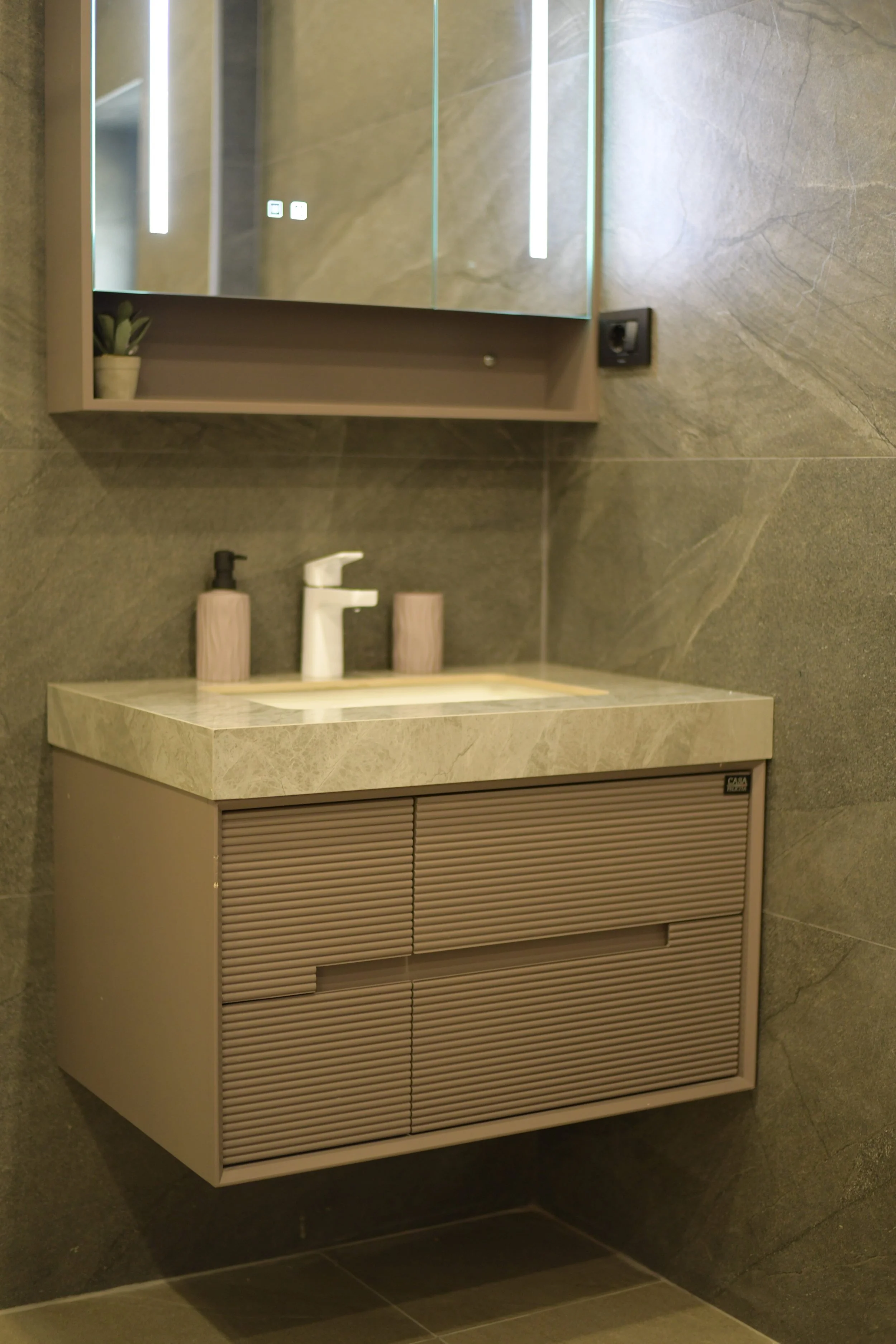 Modern bathroom vanity with a beige drawer unit and a marble countertop, above a tiled wall with a mirror and a small potted plant.